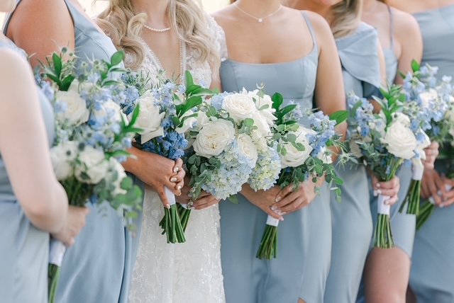 Bride and bridesmaids holding bouquets of white and blue flowers at a wedding.