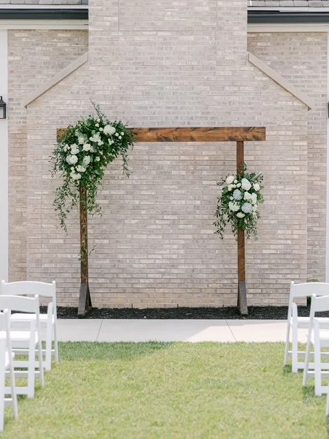 Wedding arch decorated with white flowers and greenery, set up outdoors on grass with white chairs nearby.