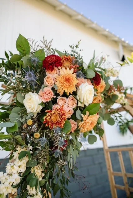 Close-up of a colorful floral arrangement with peach, red, white, and orange flowers and green leaves, set outdoors against a house with a white wall and blue sky.