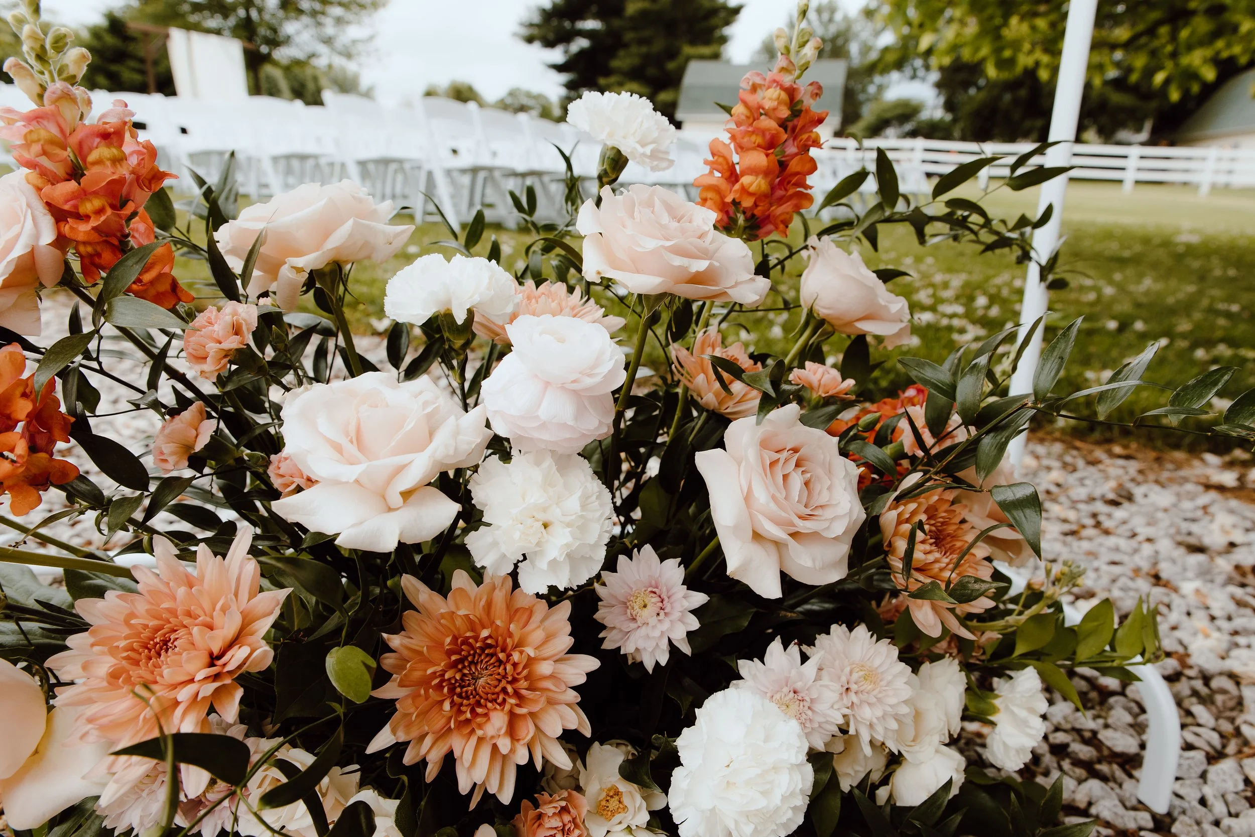 Close-up of a floral arrangement with peach, white, and orange flowers outdoors, with chairs and trees in the background.