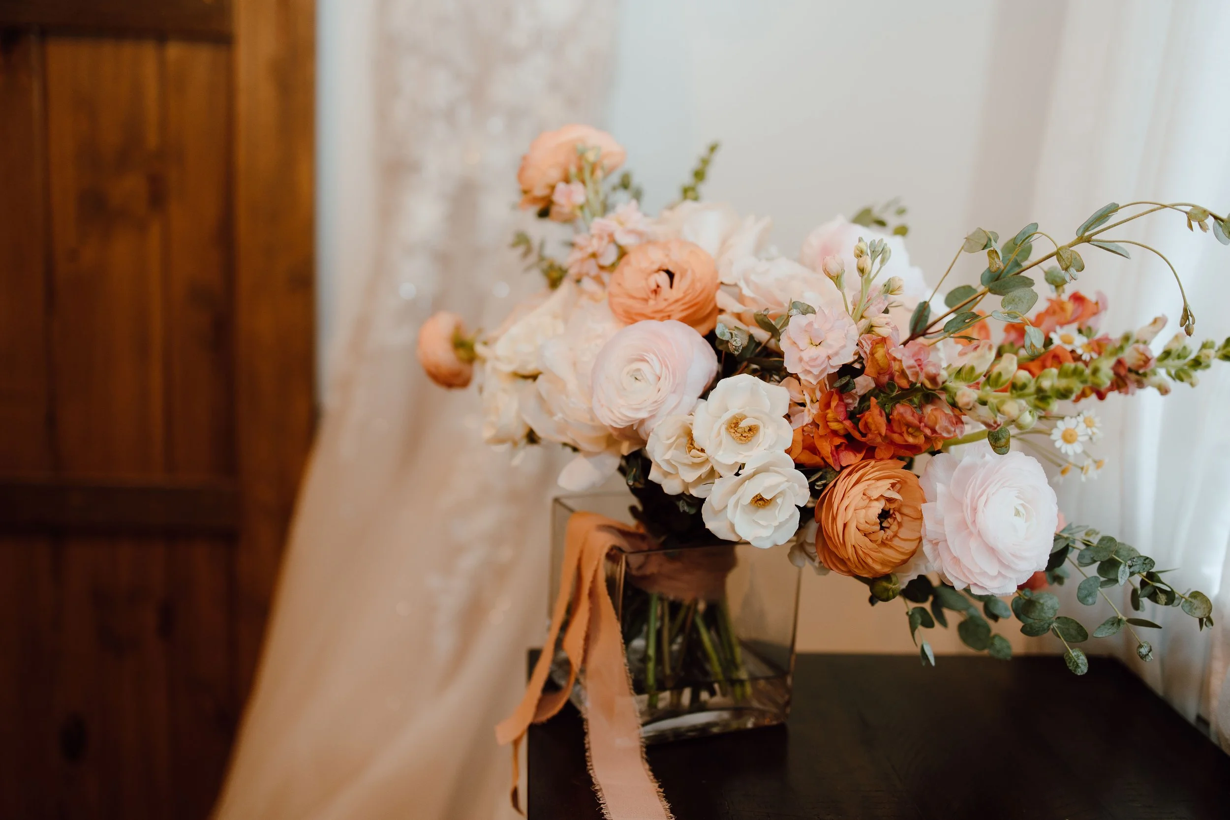 A flower arrangement with peach, white, and blush pink flowers, eucalyptus leaves, and greenery in a black vase on a table, with a ribbon tied around the vase.