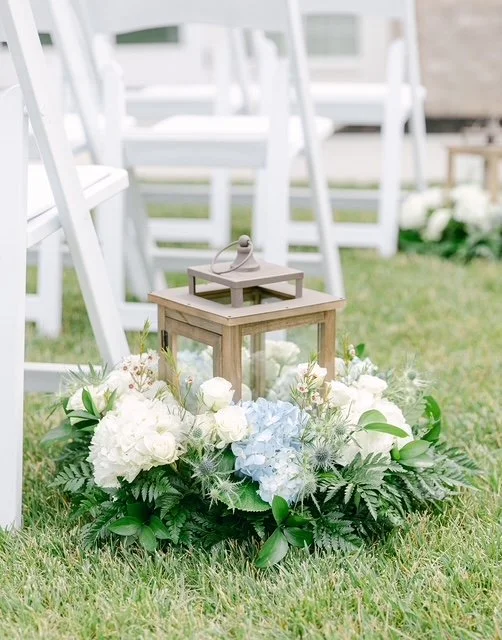 A decorative lantern surrounded by white and light blue flowers and green foliage, set on a grassy outdoor area near white chairs.