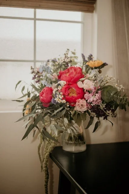 Flower bouquet with pink, red, purple, and white flowers in a clear glass vase on a black table in front of a window