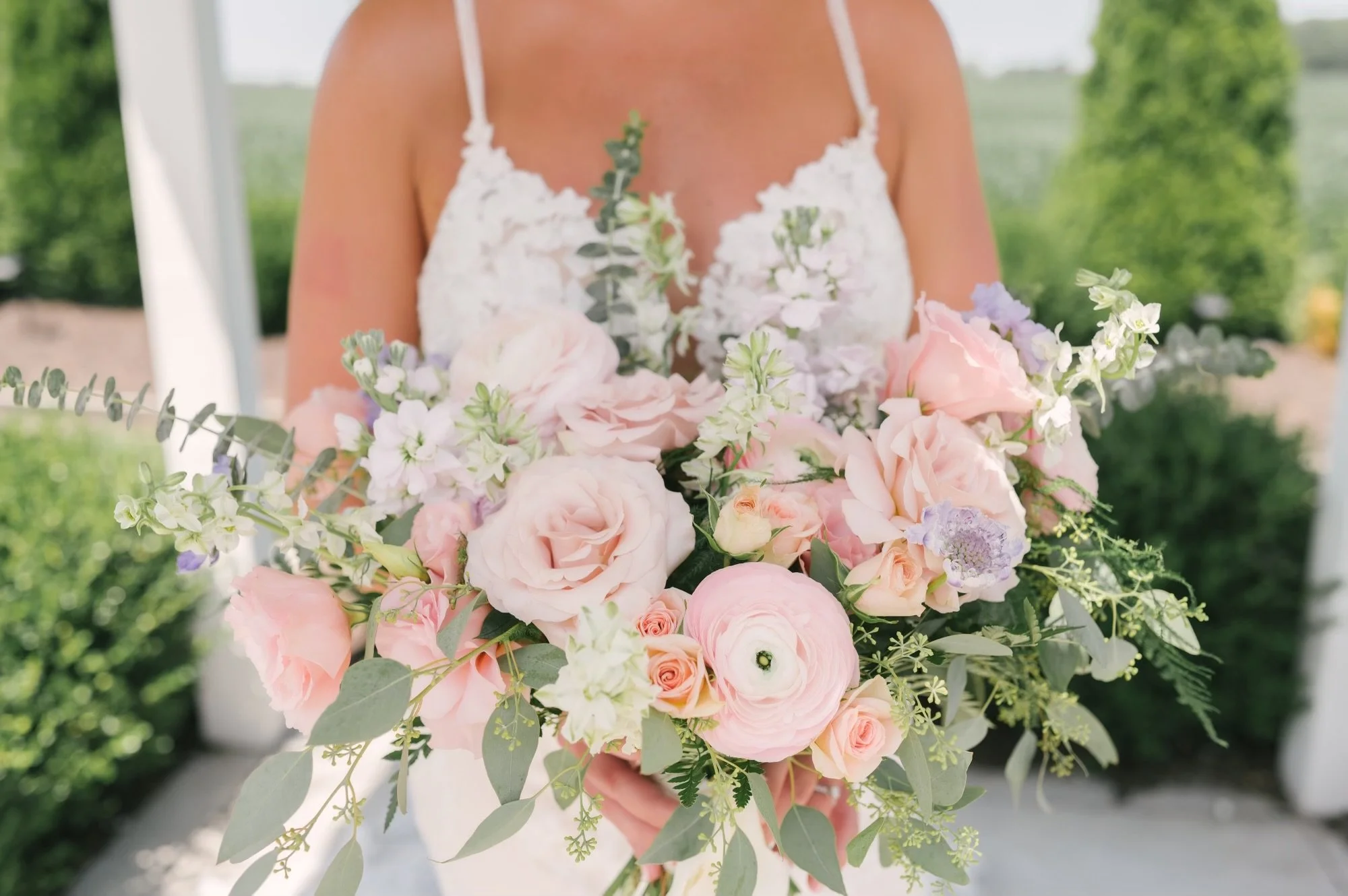 A person wearing a white dress holding a large bouquet of pink, white, and purple flowers with green leaves, outdoors near greenery and a white structure.