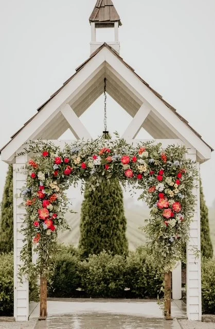 A white wooden arch decorated with red and white flowers, situated in front of a small white building with a gabled roof, surrounded by green trees and bushes.