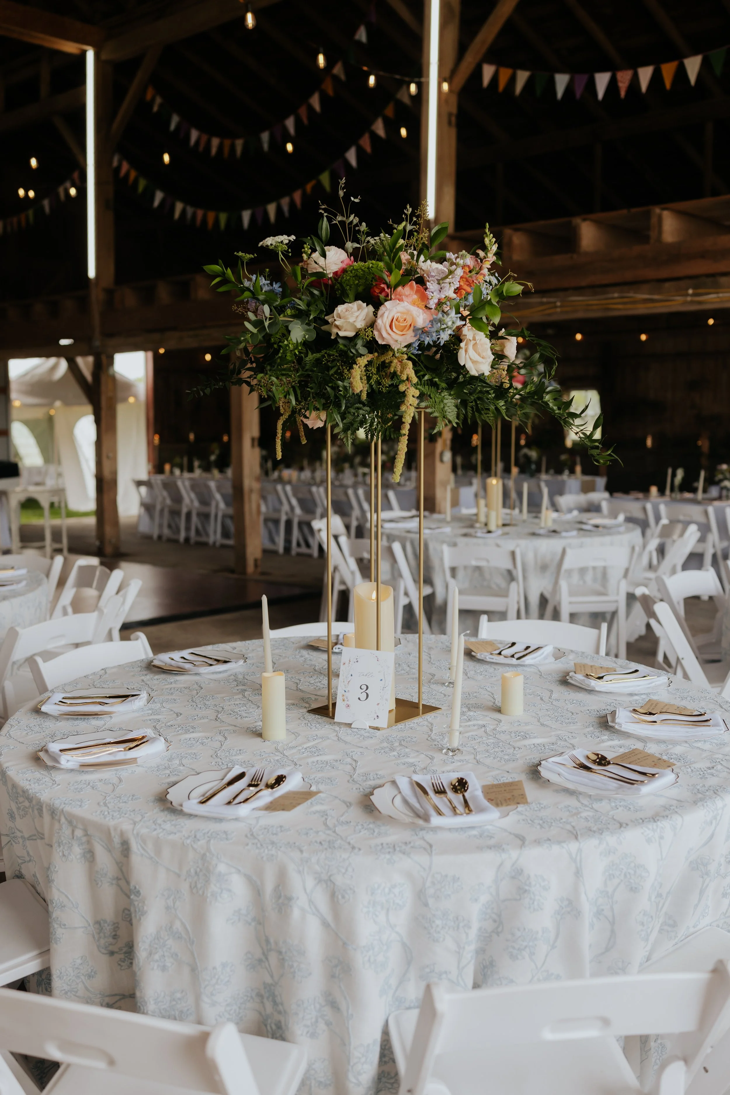 Elegant round table set for a wedding reception, decorated with a lace tablecloth, white napkins, gold utensils, and wine glasses, featuring a tall floral centerpiece with pink roses, blue, green, and white flowers, and candles, in a rustic barn with