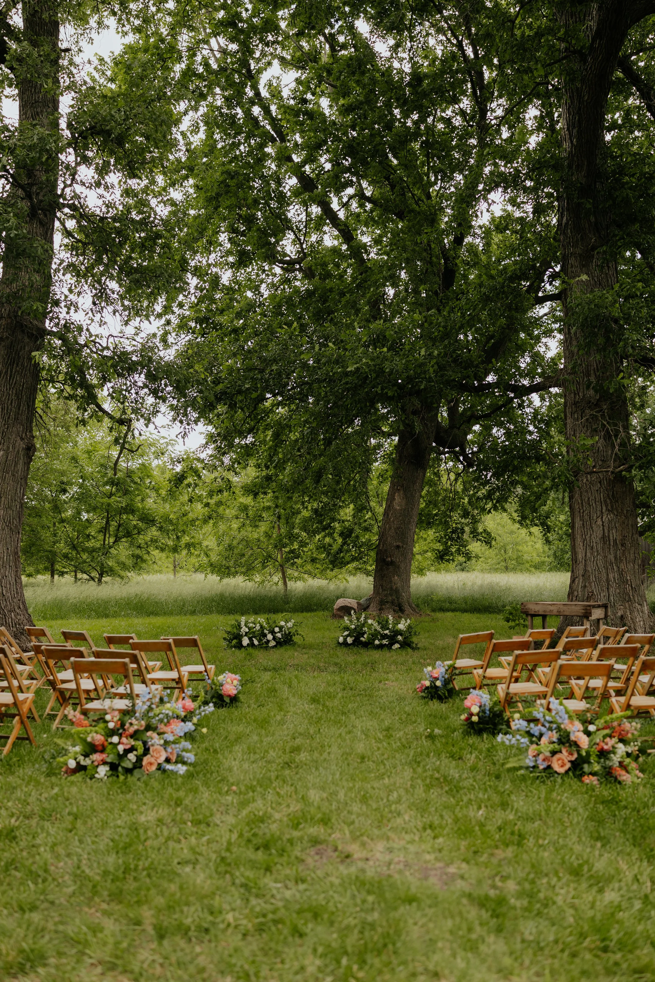 Outdoor wedding ceremony setup with wooden chairs, floral arrangements, and large trees in a grassy area.