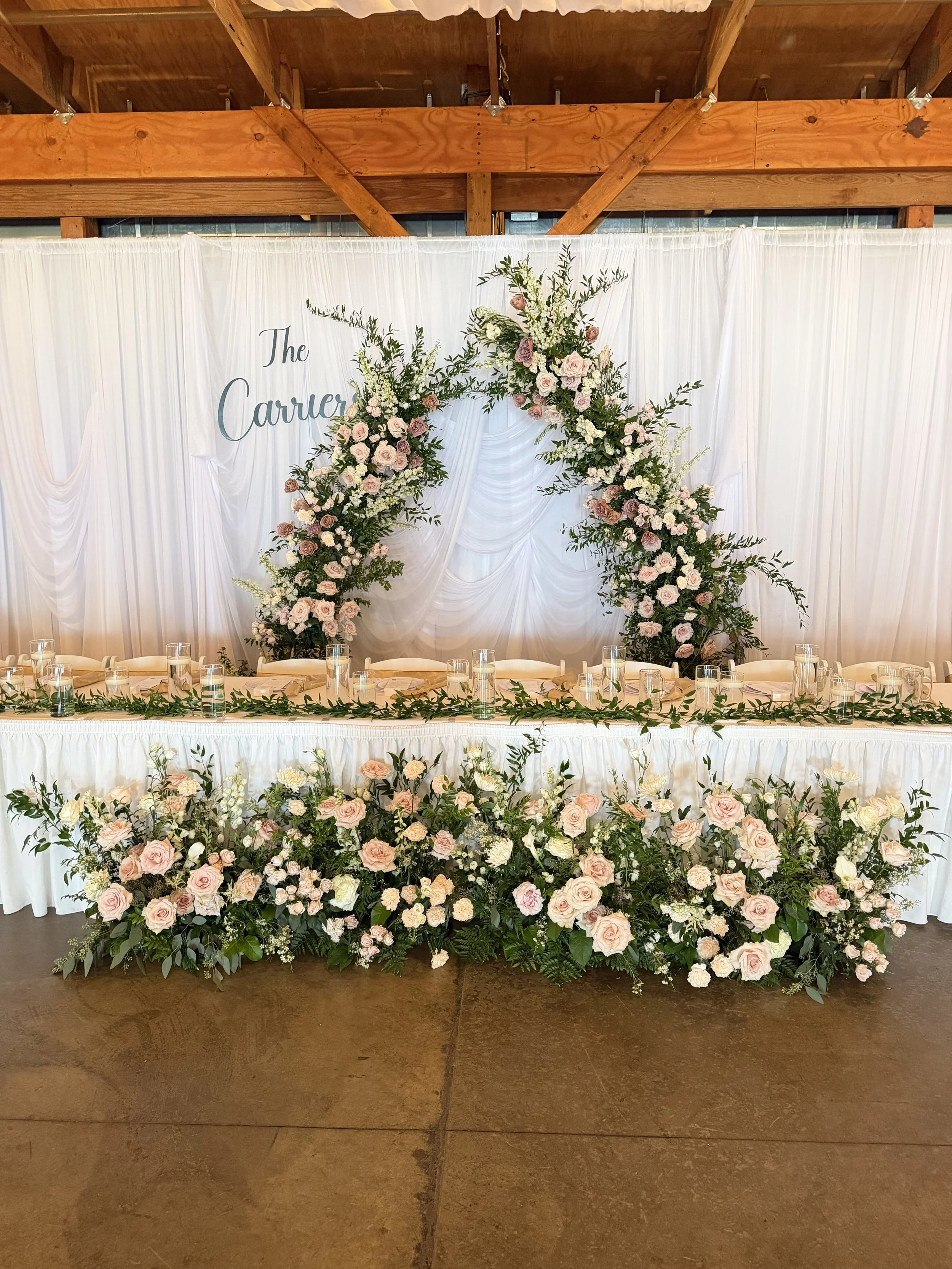 A decorated wedding or event reception table with a backdrop of white draped fabric, floral arrangements, and candles.