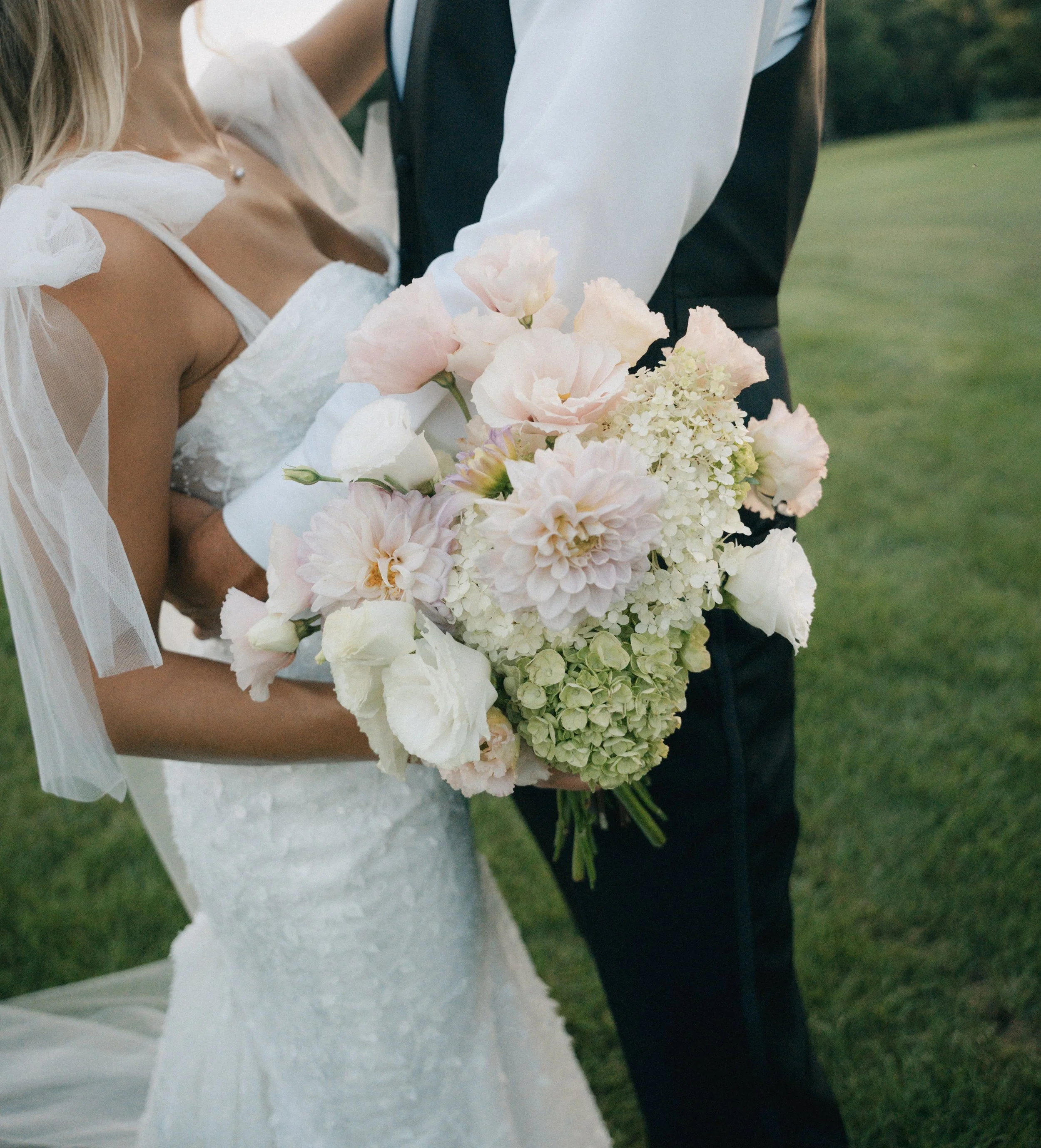 A bride holding a bouquet of pale pink and white flowers while standing with the groom on a grassy field.