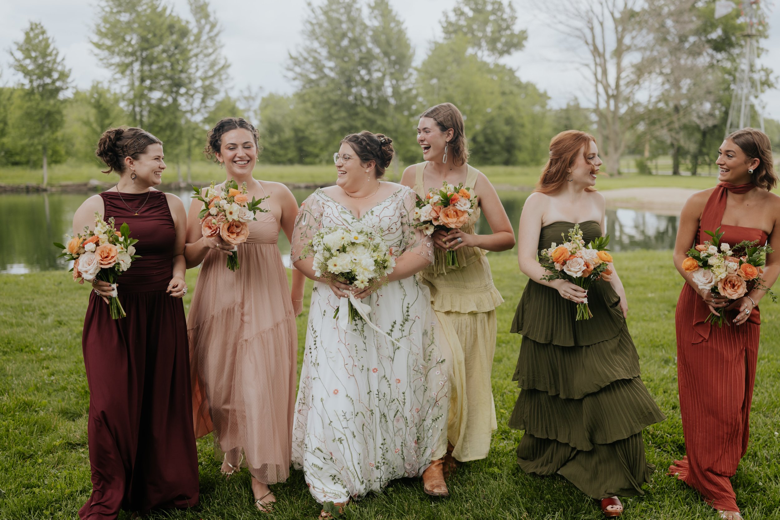 Group of six women standing outdoors on grass near a pond, smiling and holding bouquets of flowers, dressed in colorful dresses for a wedding or celebration.