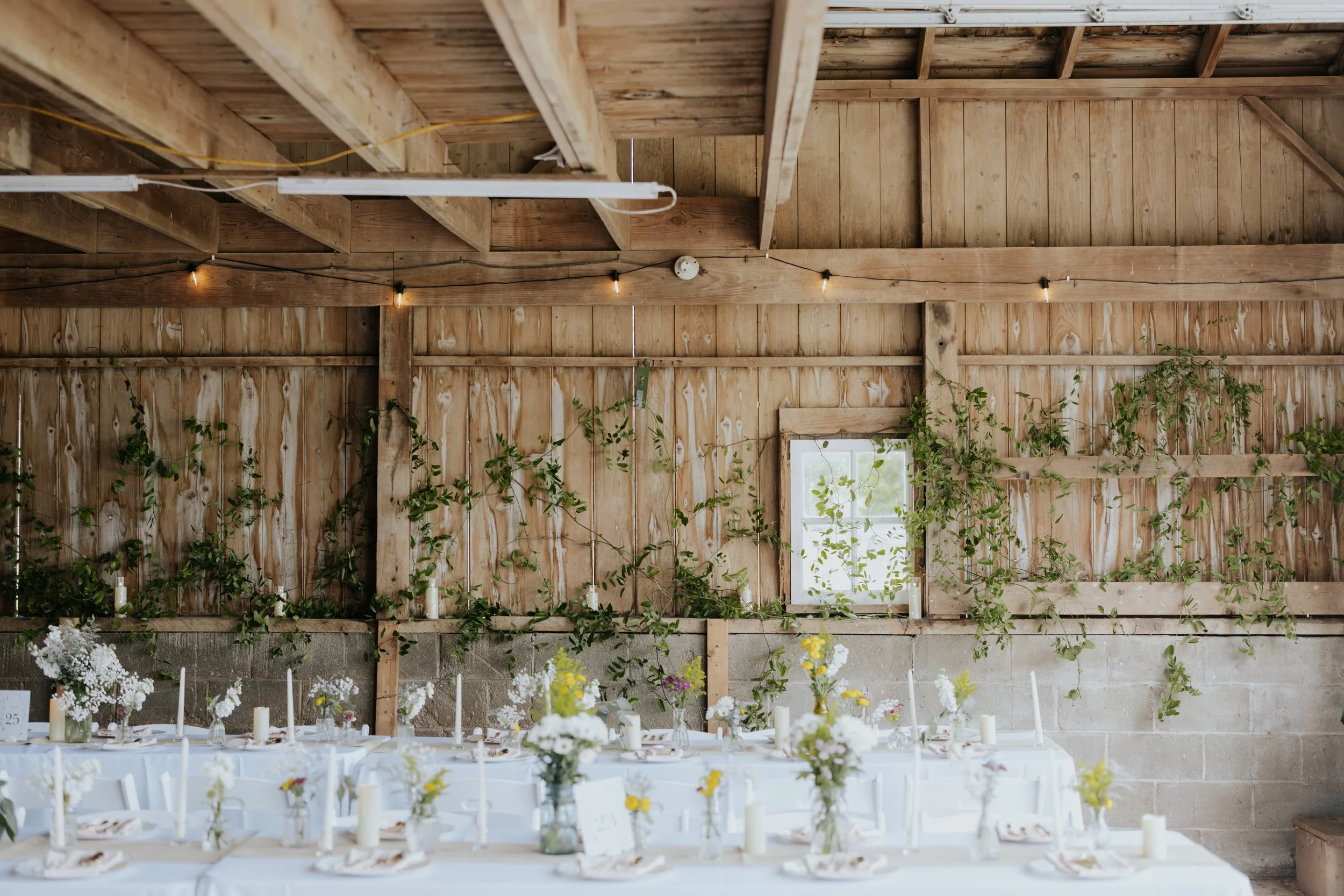 Interior of a rustic wooden barn decorated for a wedding or celebration with tables set up with white tablecloths, candles, and floral arrangements, and string lights hanging from the ceiling.