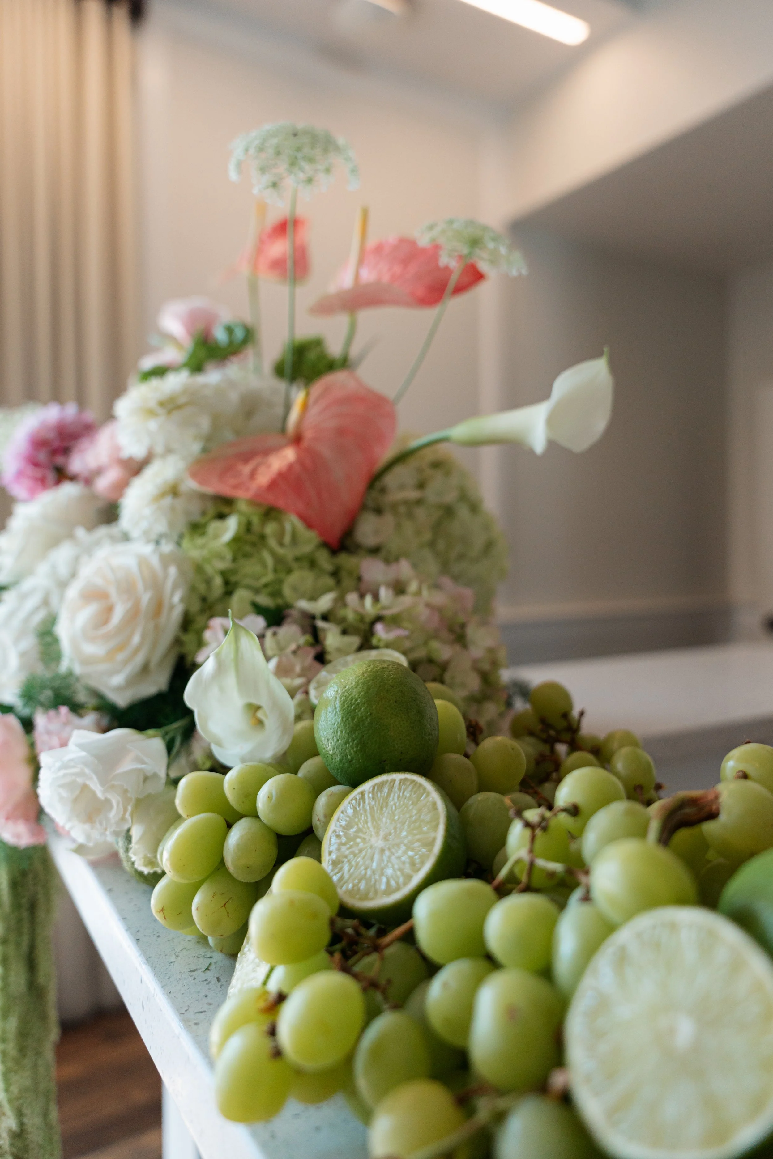 Close-up of a floral arrangement featuring white roses, pink and white anthuriums, green grapes, and slices of lime and lemon, with a blurred background of a room interior.