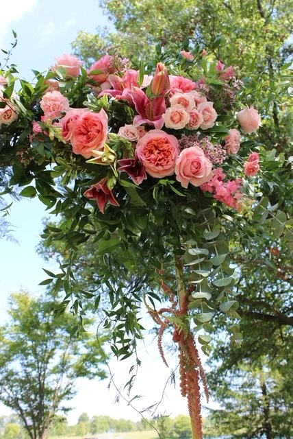 A tall bouquet of pink roses, lilies, and other pink flowers with green foliage, set outdoors under a blue sky with trees in the background.