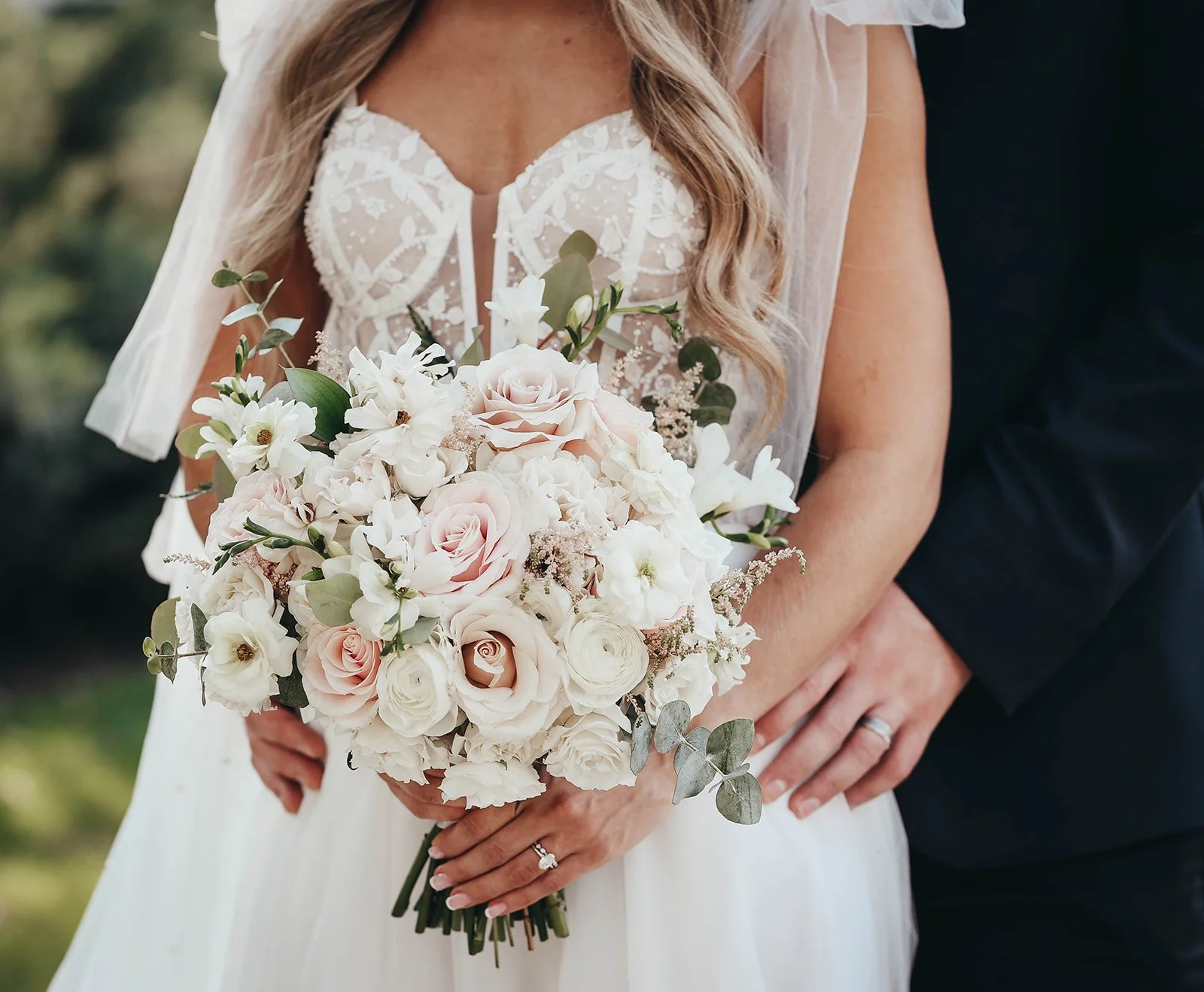 A bride holding a bouquet of roses, lilies, and eucalyptus, with her left hand resting on her lower abdomen and a wedding ring visible on her finger, while a groom's arm is wrapped around her waist.