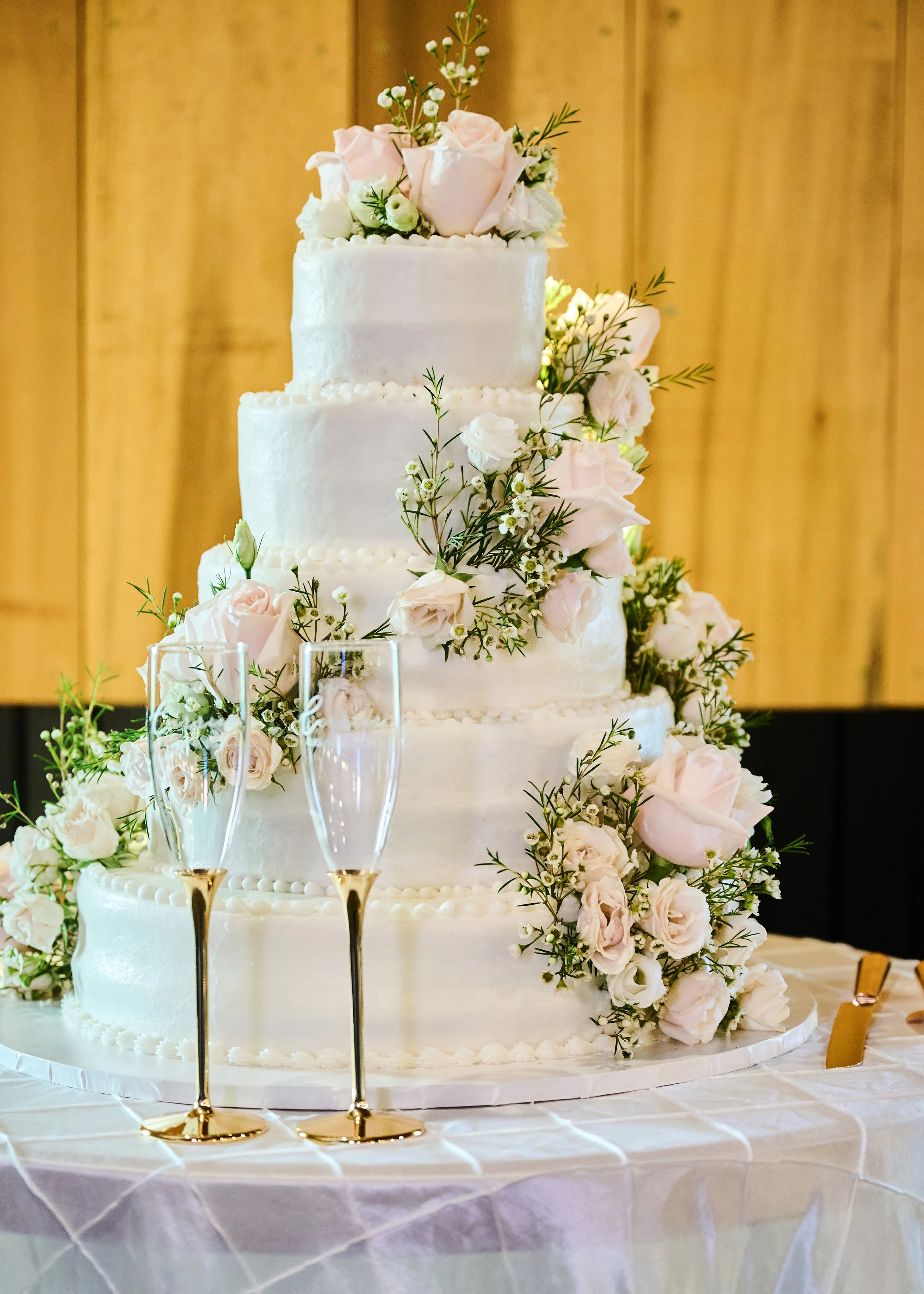 A tall, multi-tiered wedding cake decorated with pink and white roses and greenery, set on a table with a white tablecloth. In front of the cake are two champagne flutes with gold rims. The background features wooden panels.