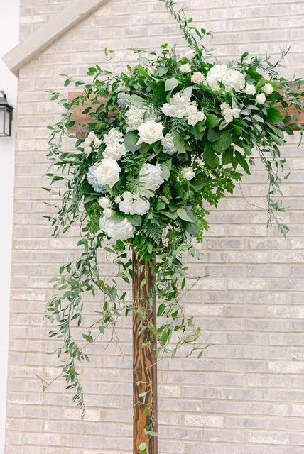 White floral arrangement with roses and hydrangeas on a tall wooden stand against a white brick wall.
