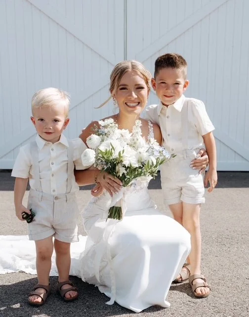 A woman dressed as a bride holding a bouquet of white flowers, kneeling with two young boys dressed in white shirts and shorts, outside in front of a white barn door.