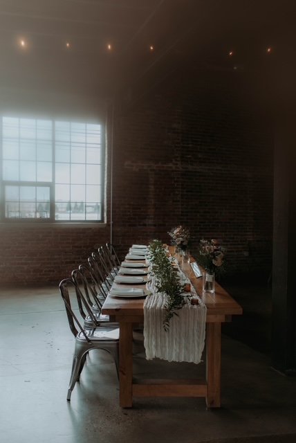 A long dining table set with plates, glasses, and floral centerpieces in a dimly lit room with a large window and brick wall.
