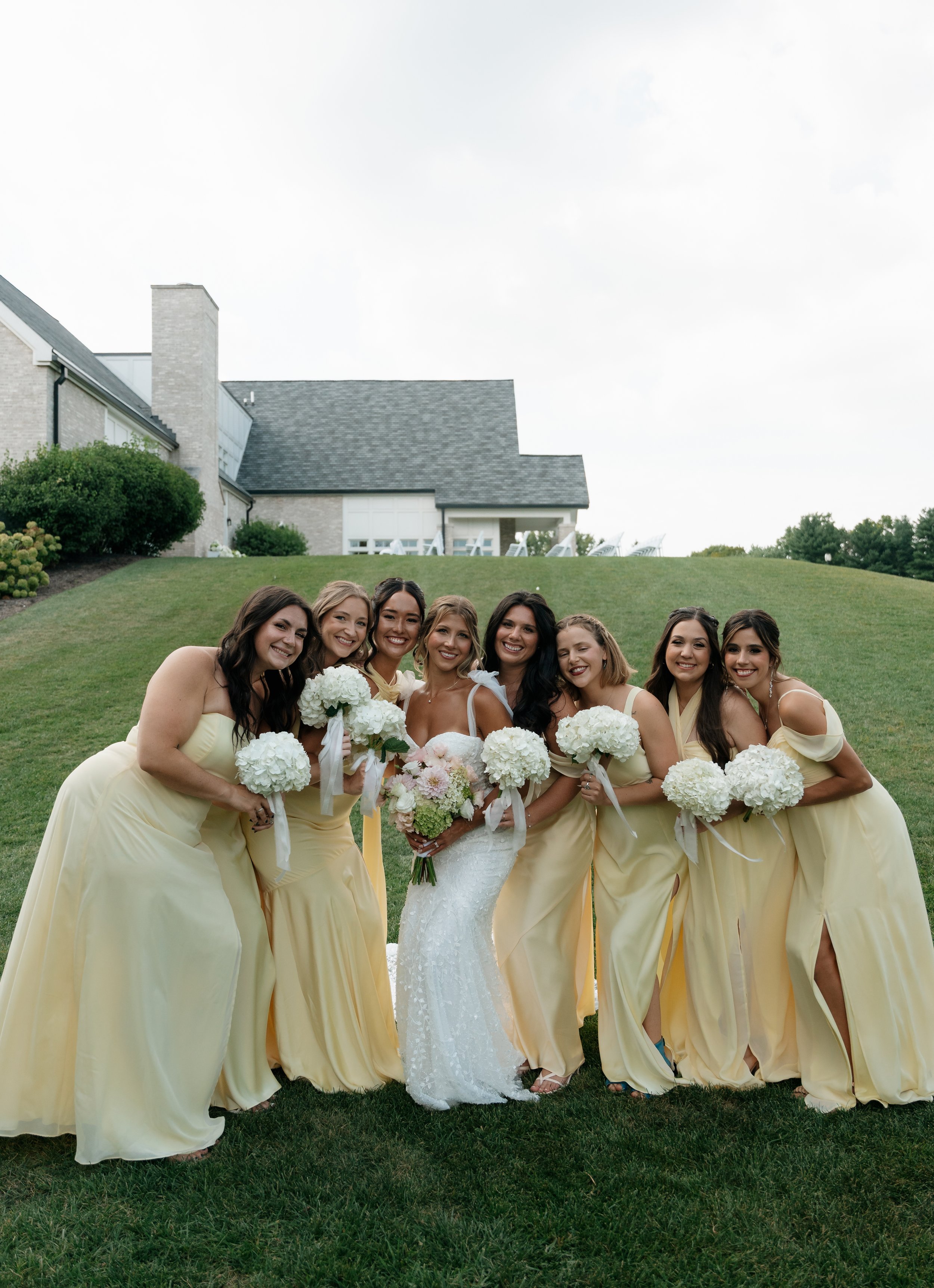 Bridal party consisting of nine women, including the bride in a white wedding dress holding a bouquet, and eight bridesmaids in long yellow dresses with white bouquets, posing together outdoors on a lawn in front of a house with a gray roof and trees