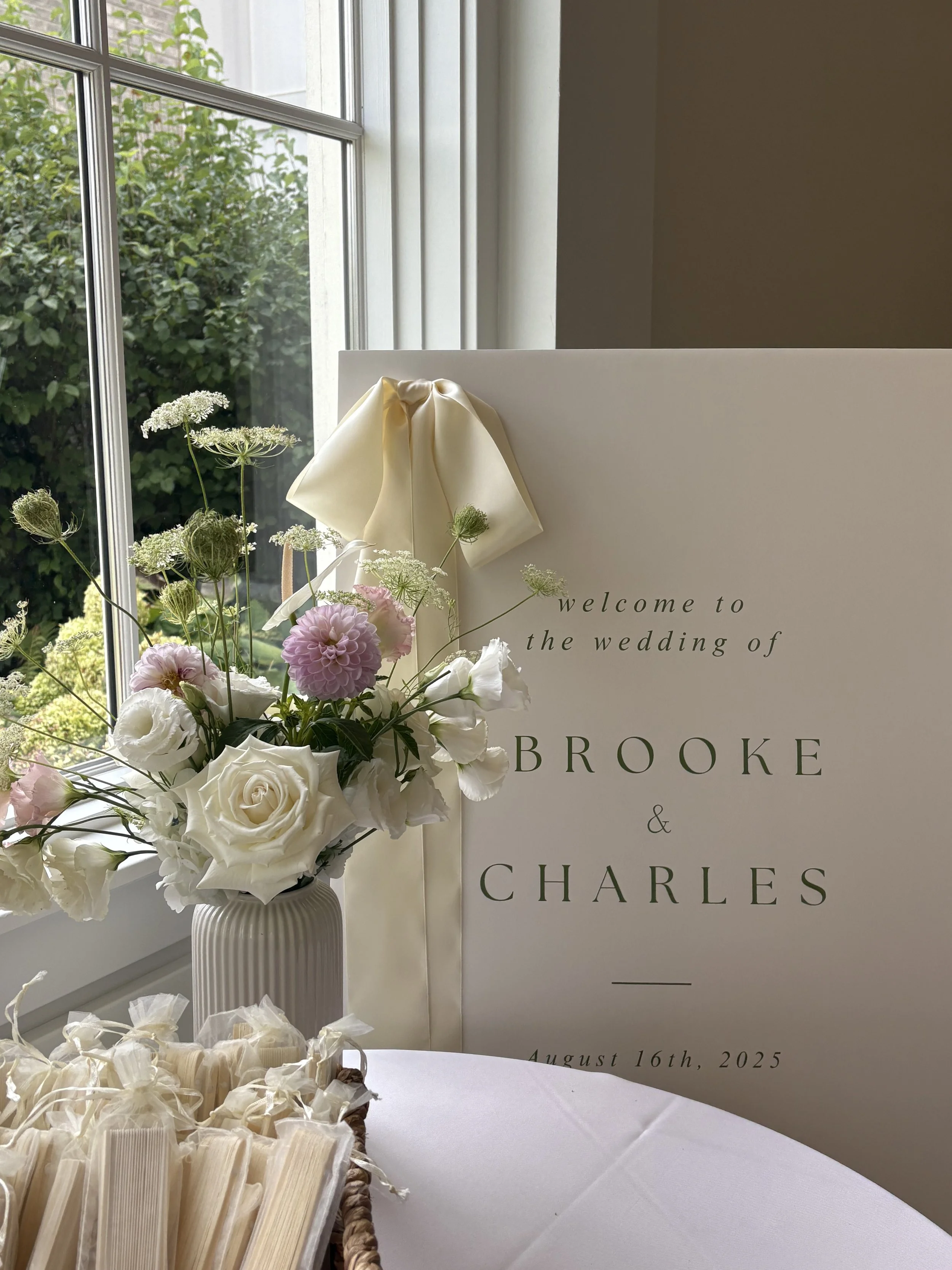 Wedding welcome sign with floral arrangement and small favor bags, near a window with greenery outside, dated August 16th, 2025.