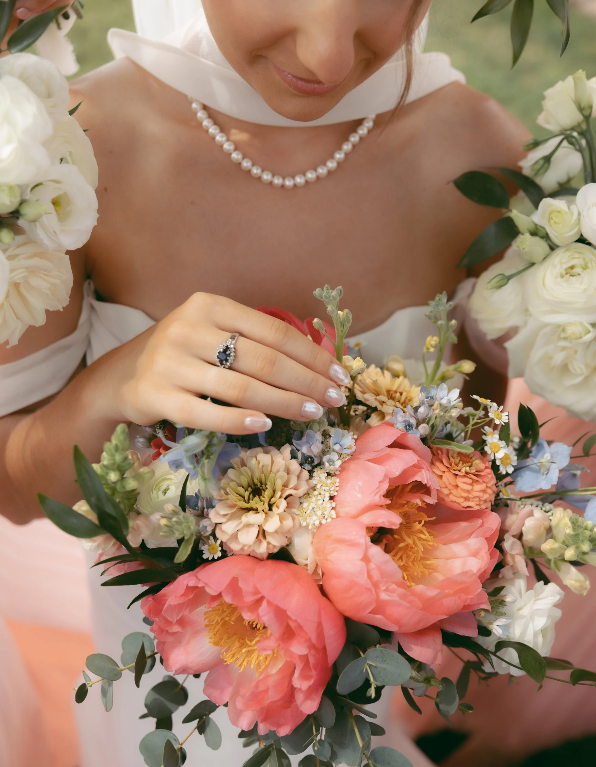 A woman in a white dress and pearl necklace holding a bouquet of colorful flowers, including pink peonies, orange dahlias, and various smaller blossoms.