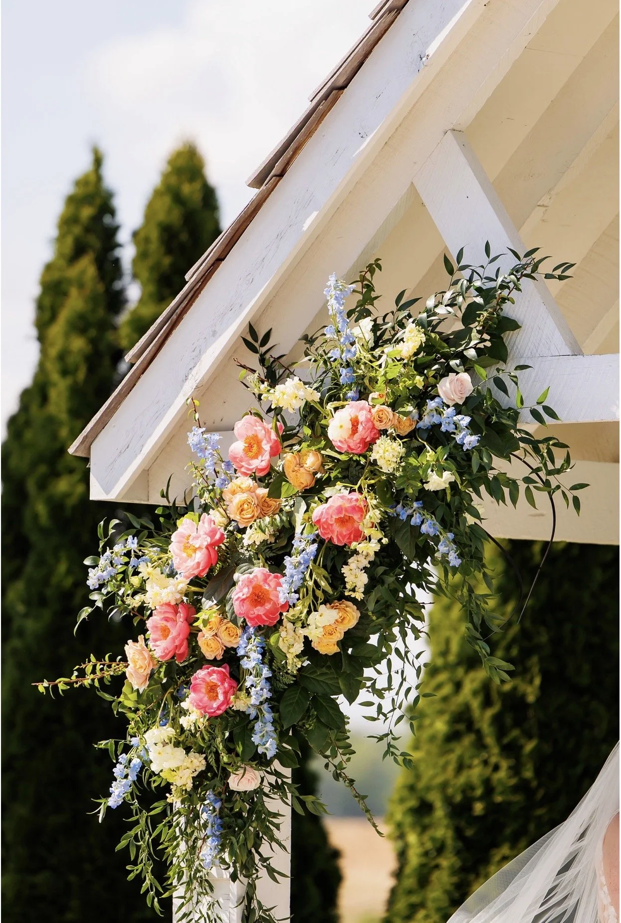 A colorful flower arrangement with pink, yellow, orange, and blue flowers, hanging from a white wooden structure outdoors.