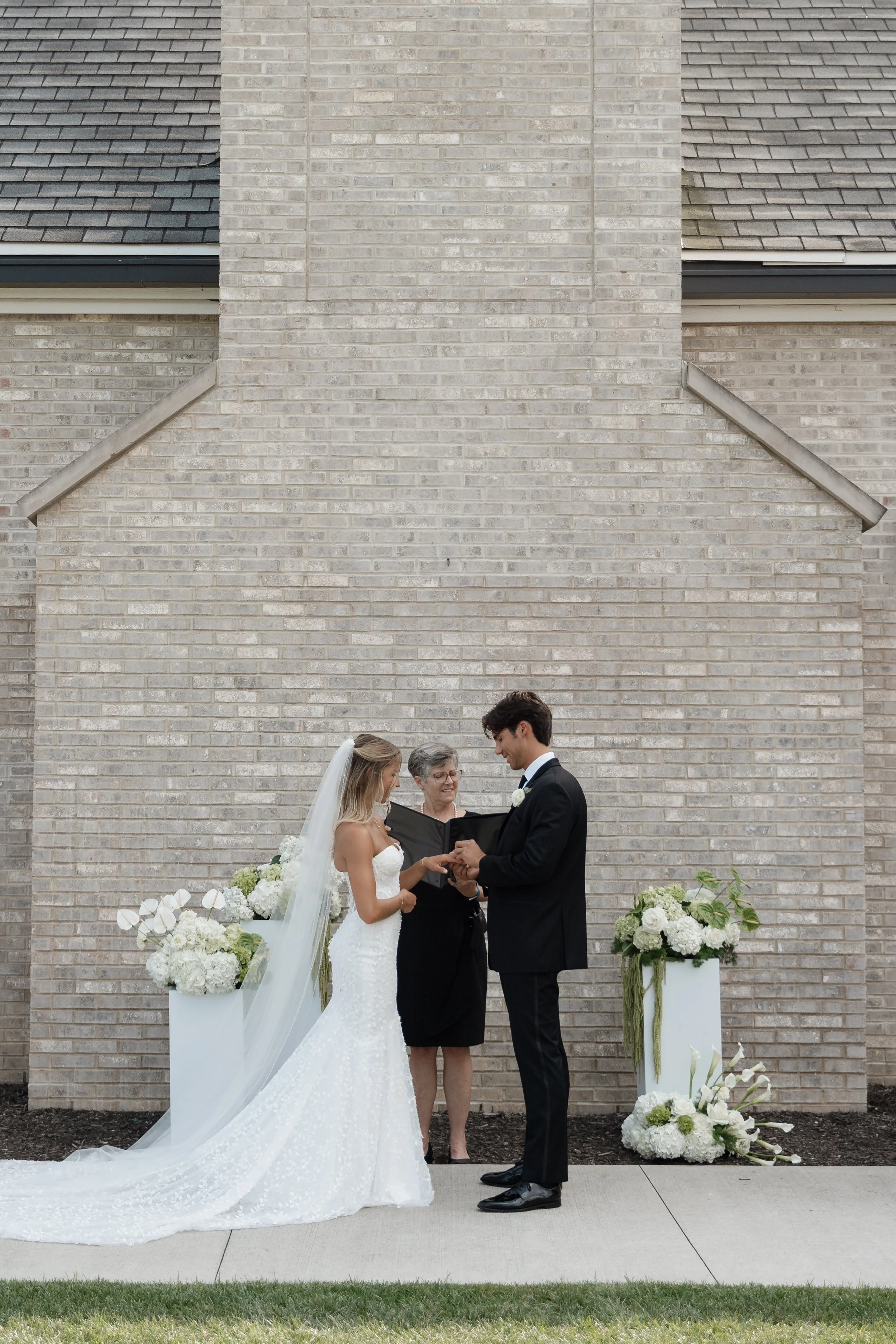 A bride and groom stand before an officiant during their outdoor wedding ceremony against a brick wall. The bride wears a white wedding gown and veil, and the groom is dressed in a black tuxedo. They are exchanging rings, surrounded by white floral a