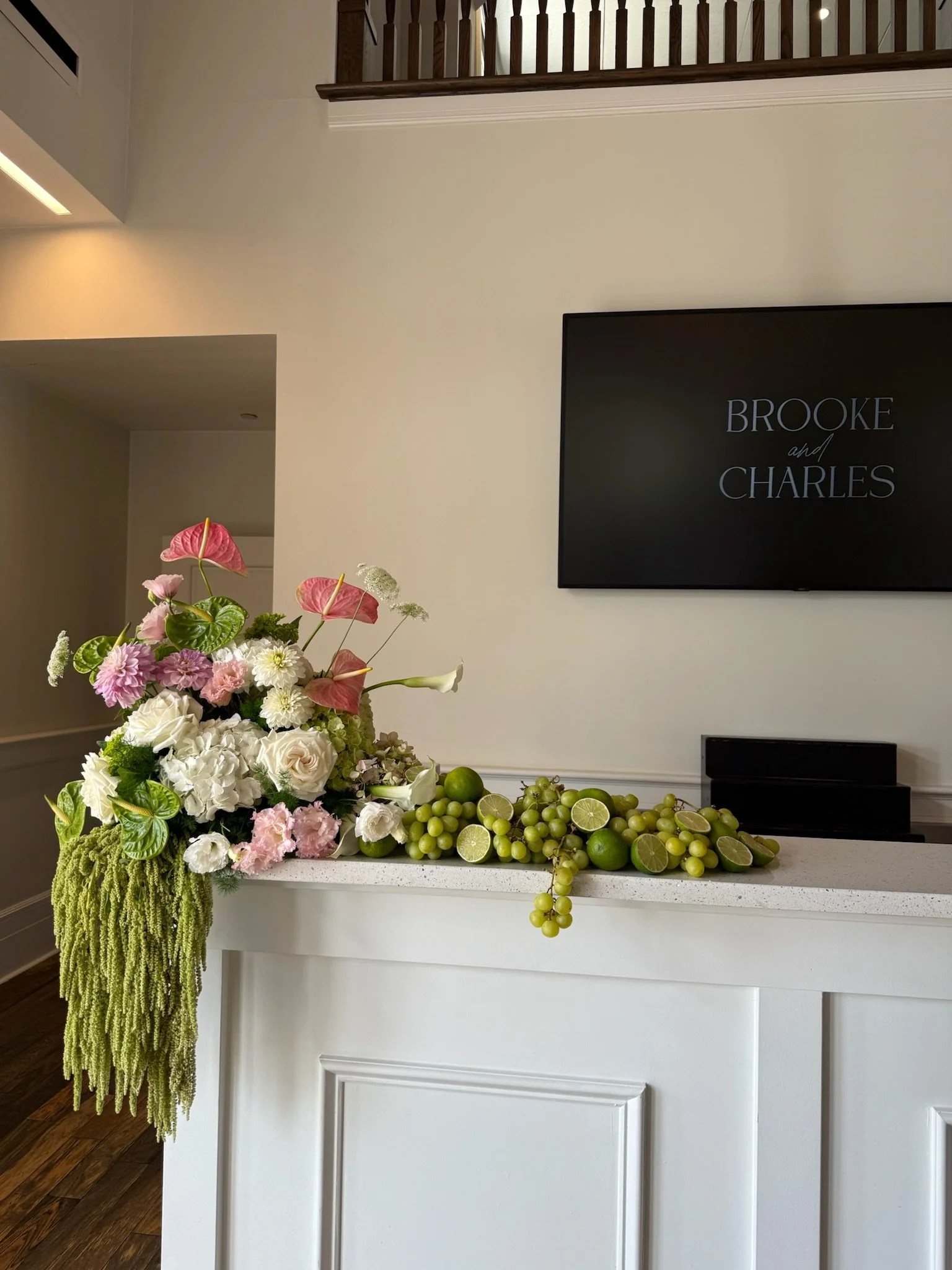 A floral arrangement with pink anthuriums, white flowers, and green foliage, along with fresh green grapes and sliced limes, on a white countertop in front of a beige wall with a black TV screen displaying "Brooke and Charles."