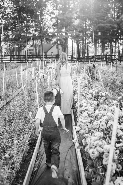 A woman leading two young children on a walkway through a garden filled with blooming flowers, with trees and a house in the background.