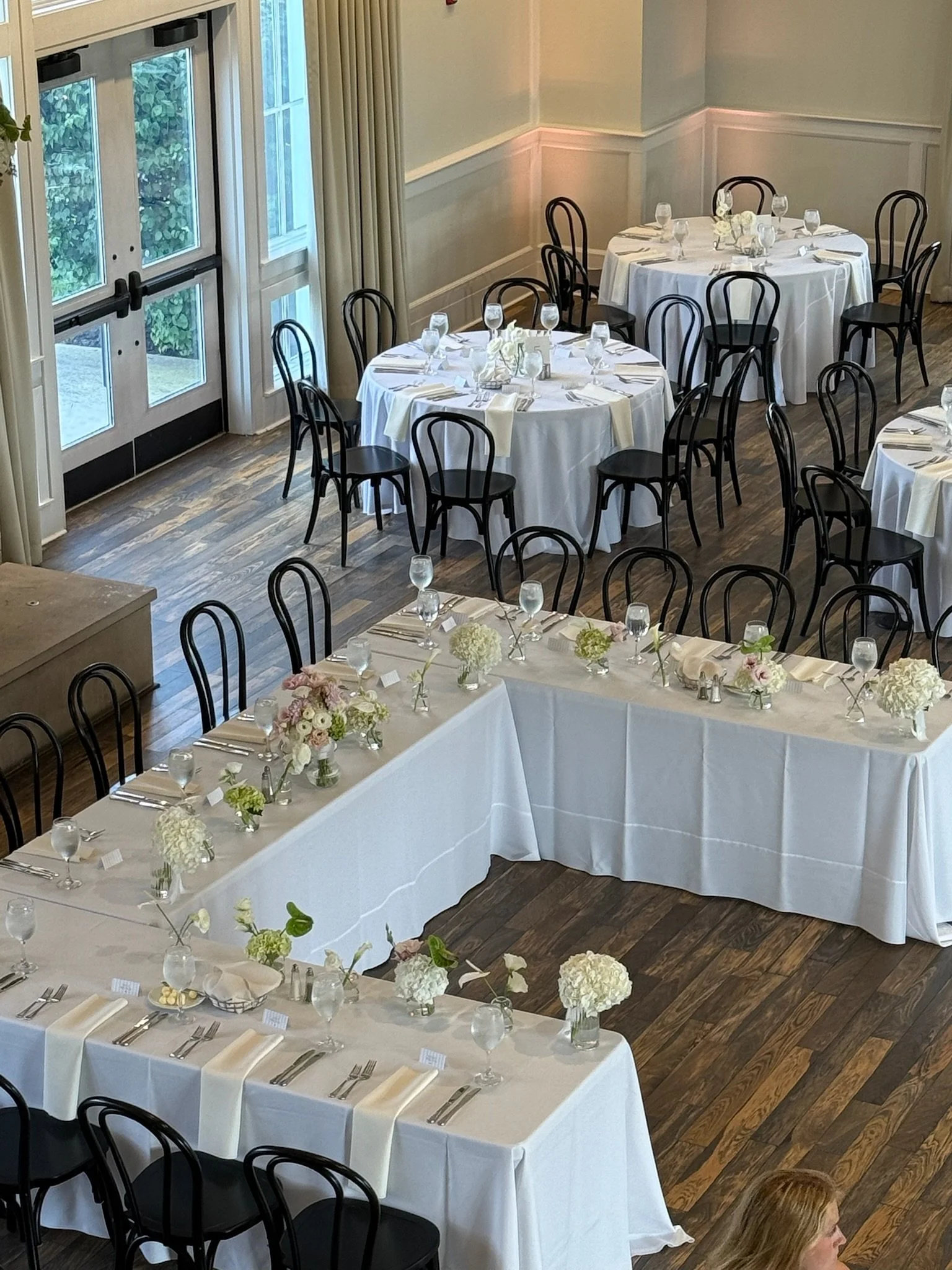 A decorated banquet room set up for a formal event with round and rectangular tables covered with white tablecloths, floral centerpieces, glassware, and place settings, with black chairs and a wooden floor.