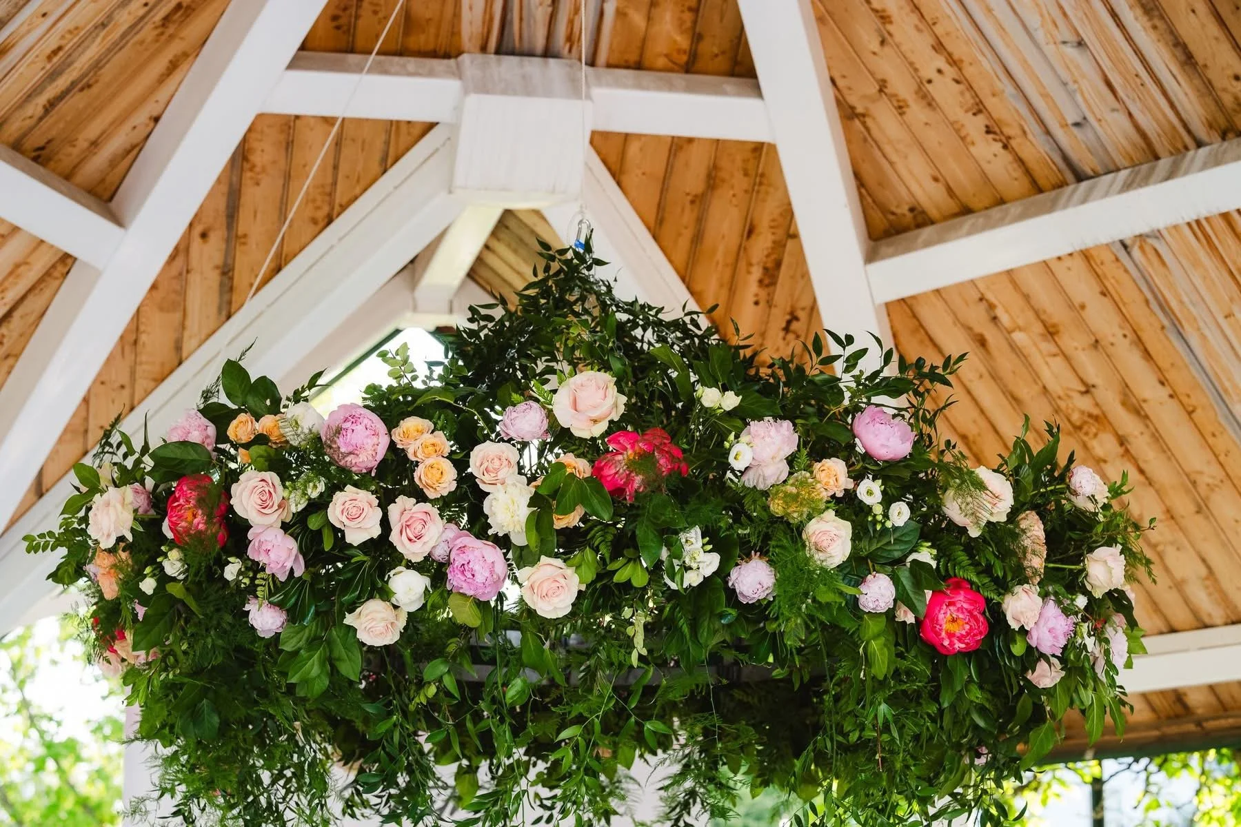 A hanging floral arrangement with pink, white, and coral roses, peonies, and greenery, suspended beneath a wooden ceiling with white beams.
