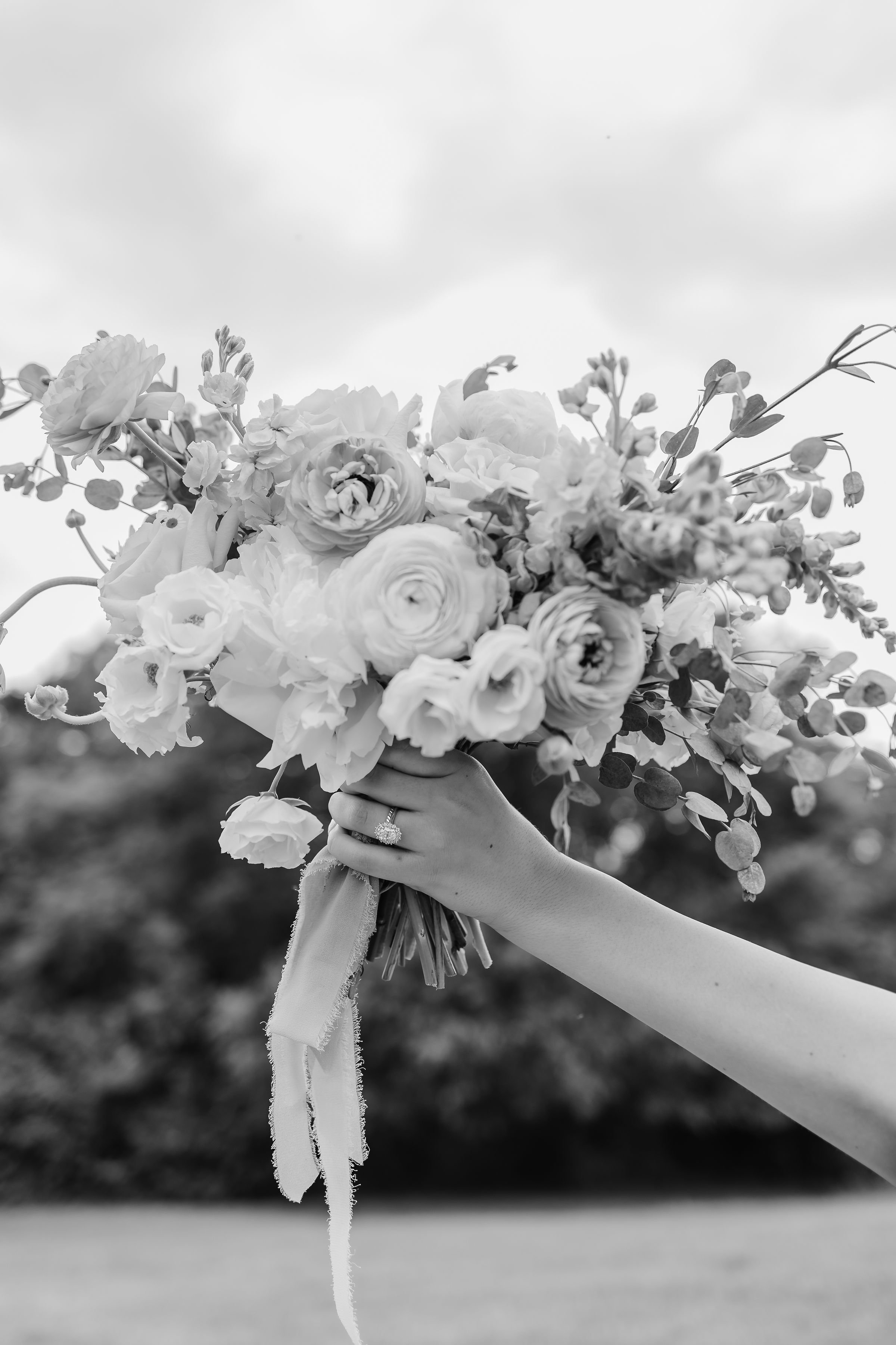 A person holding a bouquet of flowers with a ribbon, with their hand visible and a ring on their finger, against a cloudy sky background.