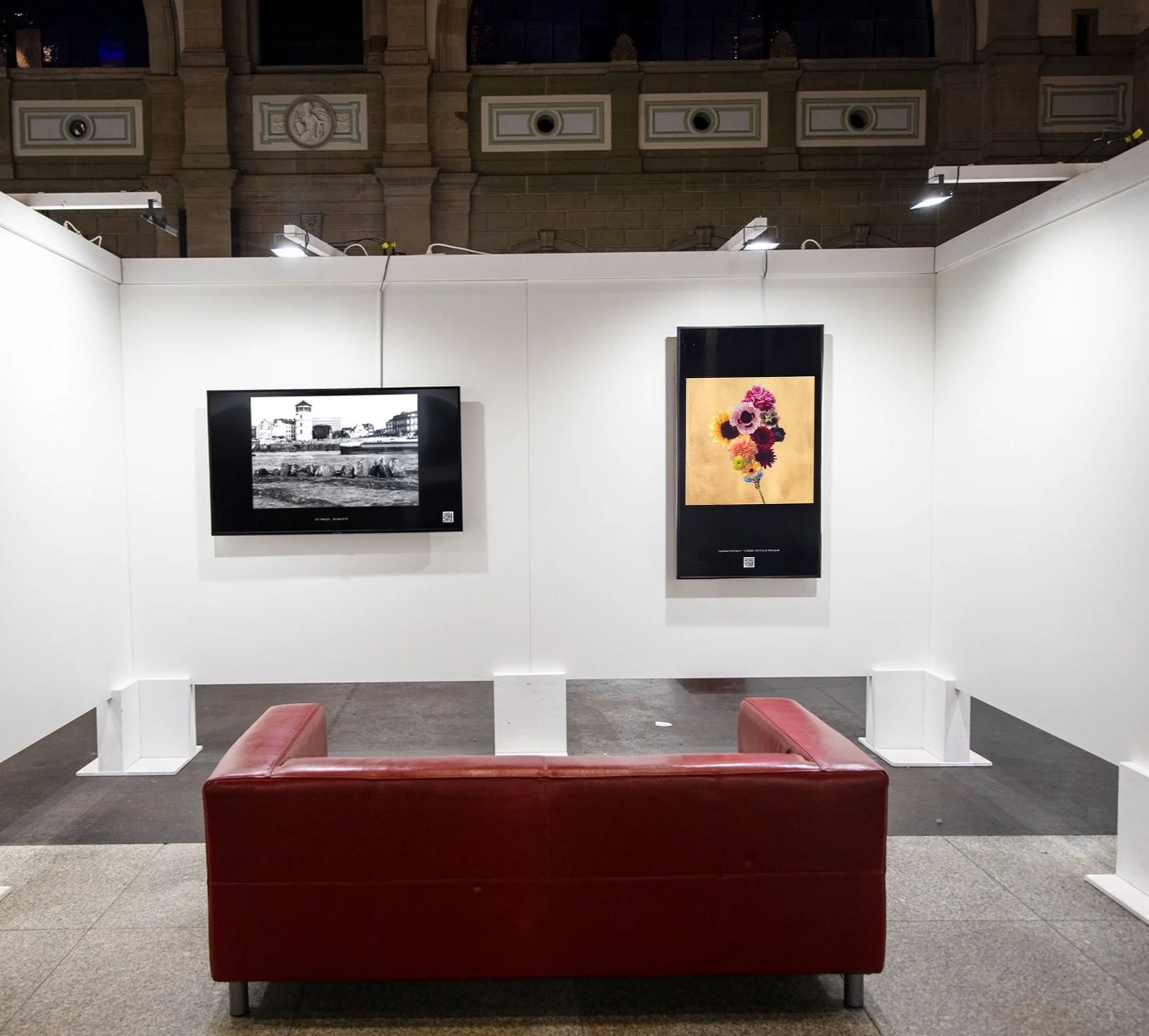 Art gallery setup with two art pieces on white walls, a television showing a black and white photo on the left, and a colorful flower artwork on the right. A red leather couch is in the foreground.