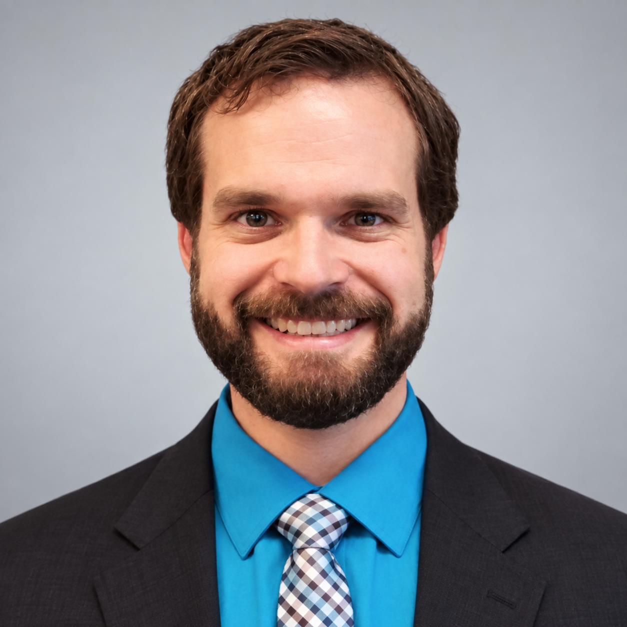 A smiling man with a beard and brown hair wearing a dark suit, blue shirt, and plaid tie, standing against a gray background.