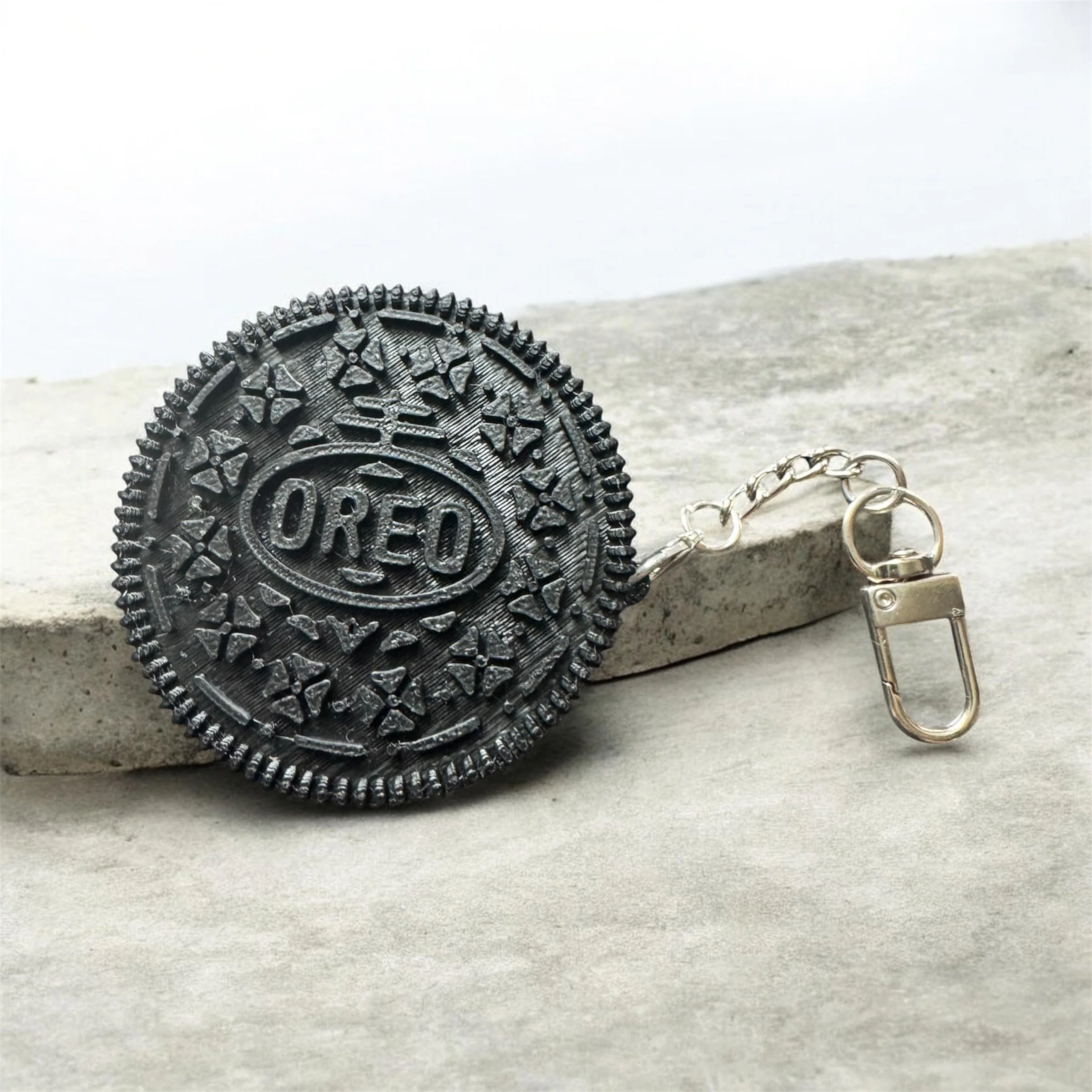 Close-up of a black Oreo cookie with a gold chain attached to a small metal clip on a light surface.