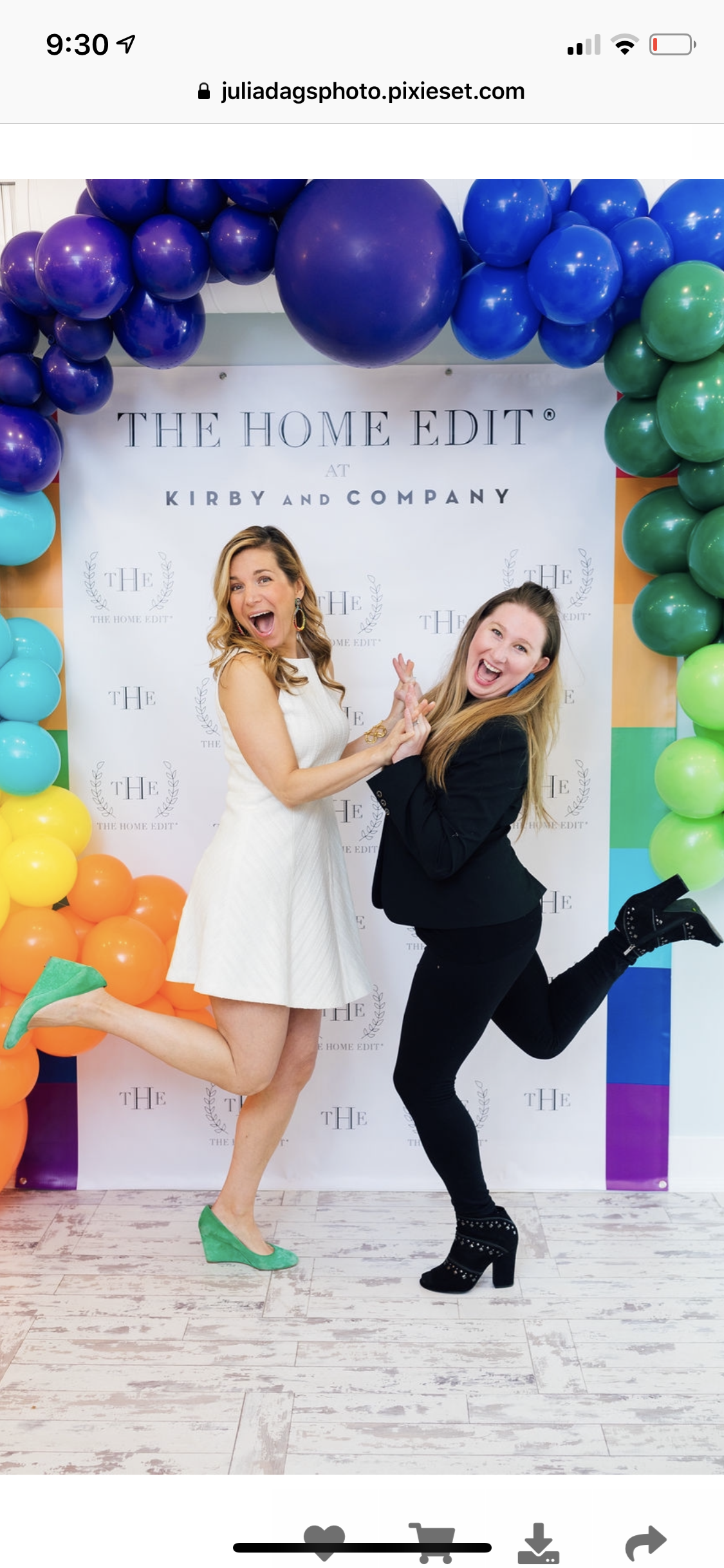 Two women jumping and smiling in front of a rainbow-colored balloon arch and a backdrop with the text 'The Home Edit at Kirby and Company'.