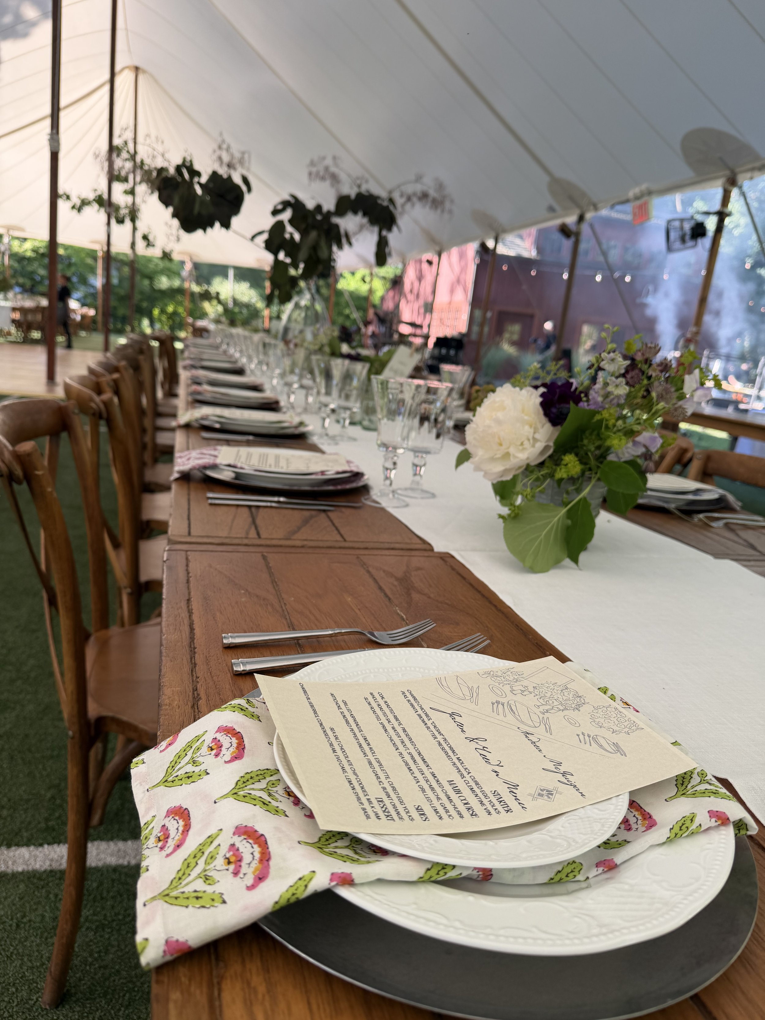 A long wooden table set for a meal under a white canopy with floral arrangements, glassware, and menus. There are wooden chairs along the table, and a floral napkin is placed on a white plate in the foreground.