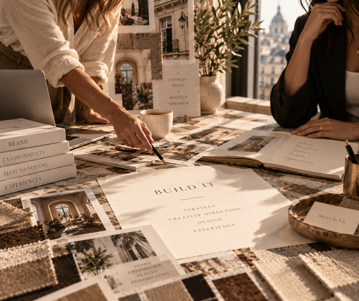 Women collaborating on a design project at a cluttered table with magazines, fabric samples, and a large paper titled 'Build It' in sunlight.