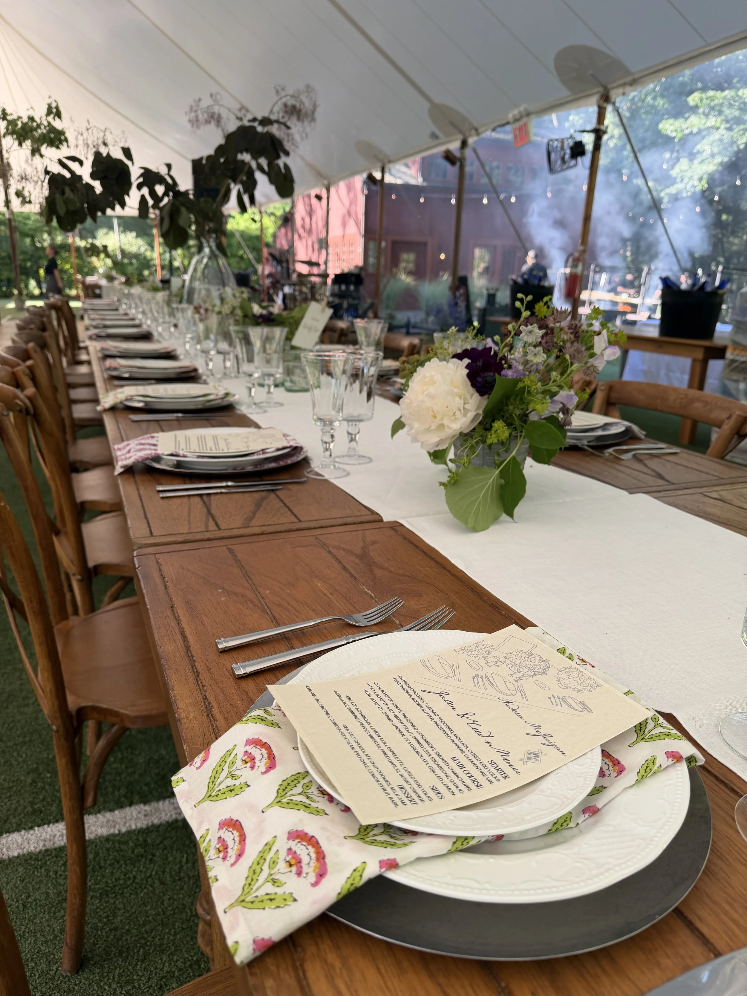 A long rectangular wooden table set for an outdoor event with glassware, plates, silverware, a floral centerpiece, and menus. The table is covered with a white runner, and a floral cloth is beneath the plates. There is a tent overhead, and in the background, there are trees, string lights, and a building with steam or smoke rising.