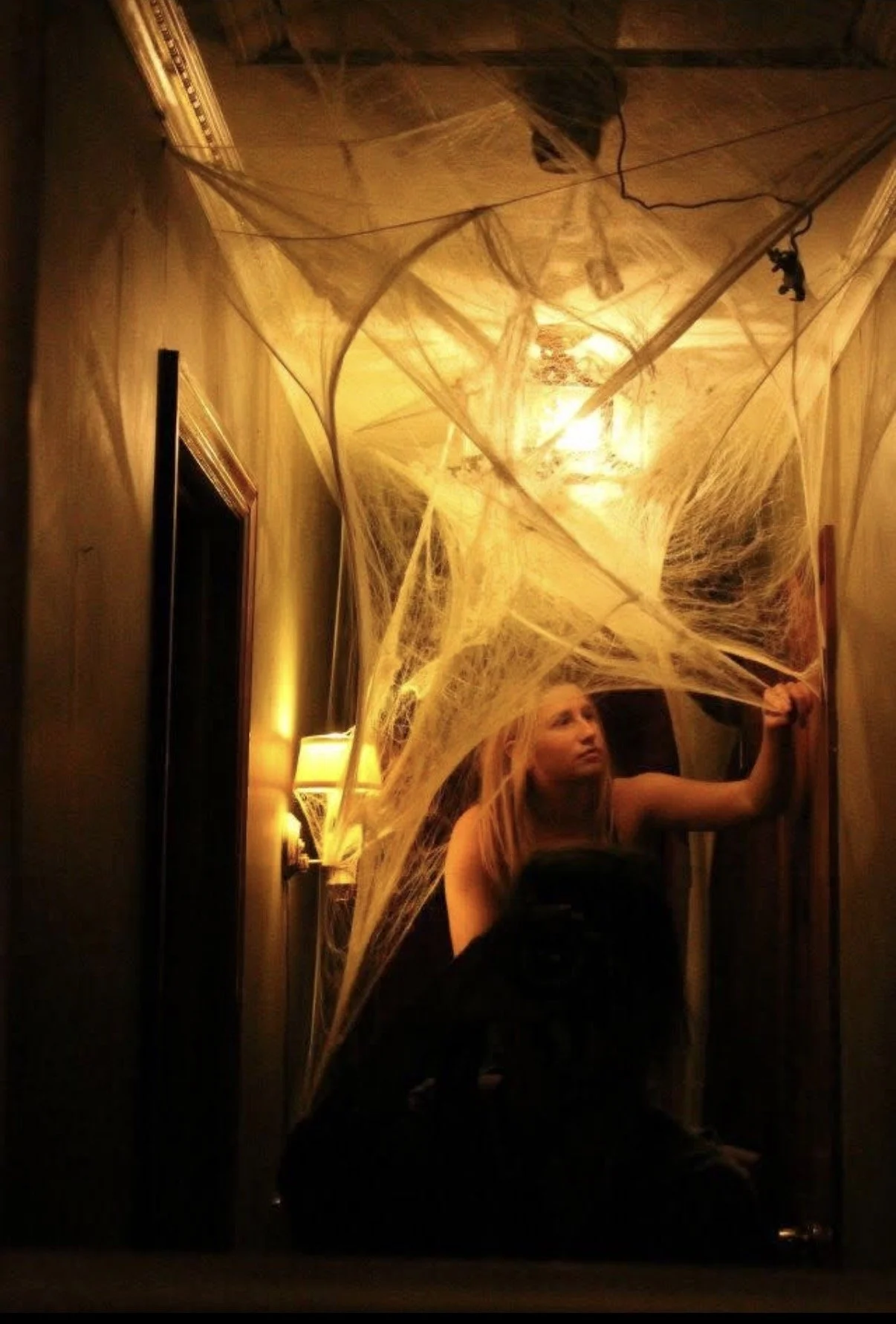 A woman sitting and holding a large web of fake spider webs in a dimly lit room, decorated for Halloween.