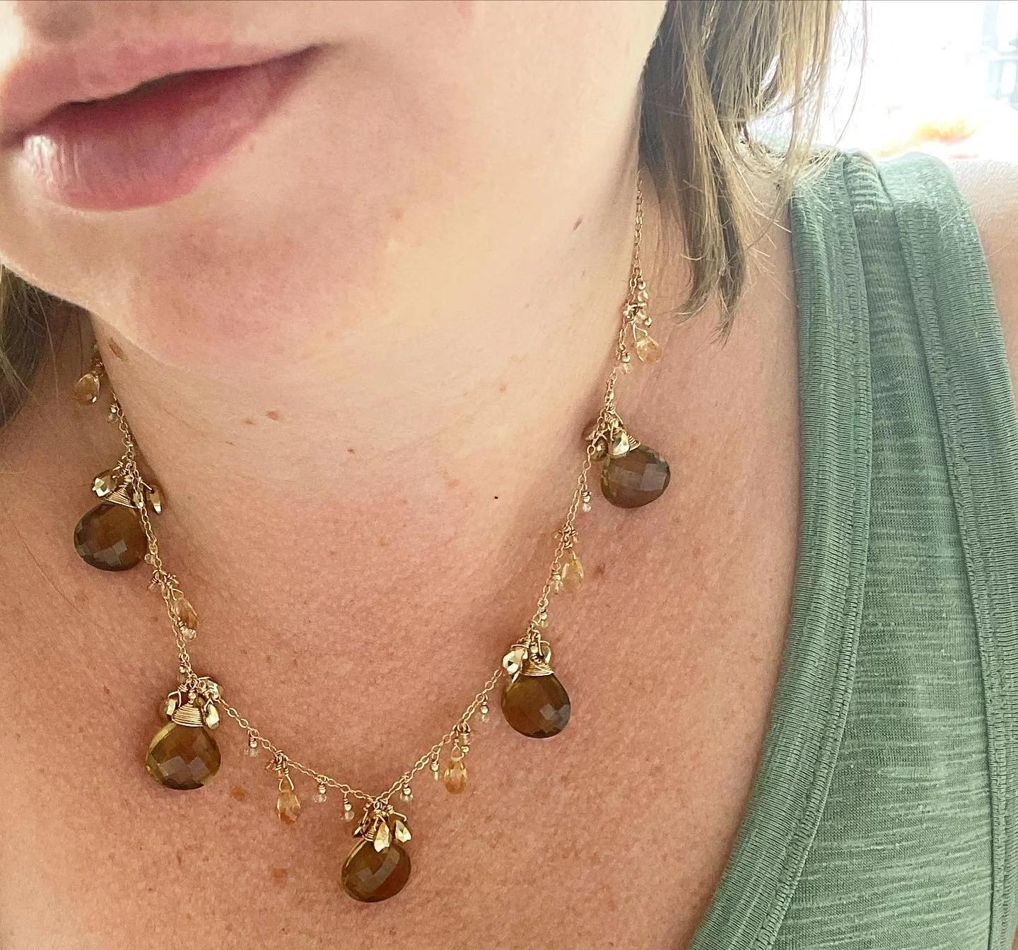 Close-up of a woman's neck and chest wearing a gold necklace with large brown gemstones and small clear beads, with a green sleeveless top visible on her shoulder.