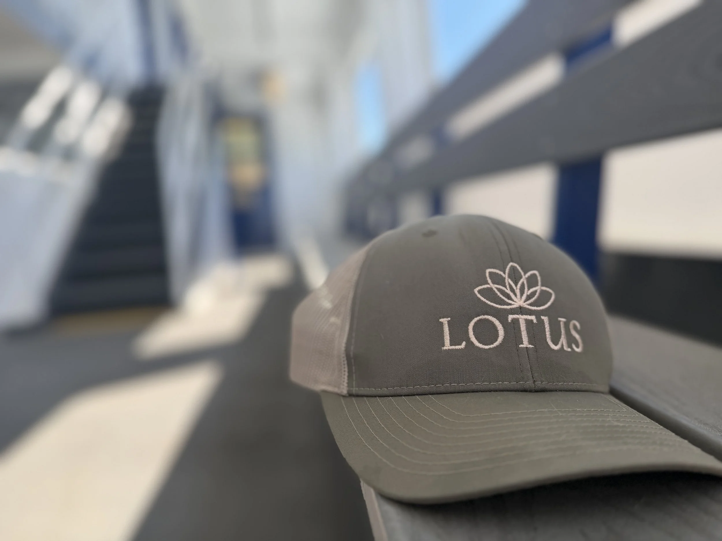Gray cap with Lotus logo resting on a metallic surface, with blurred background of a bus or train interior.