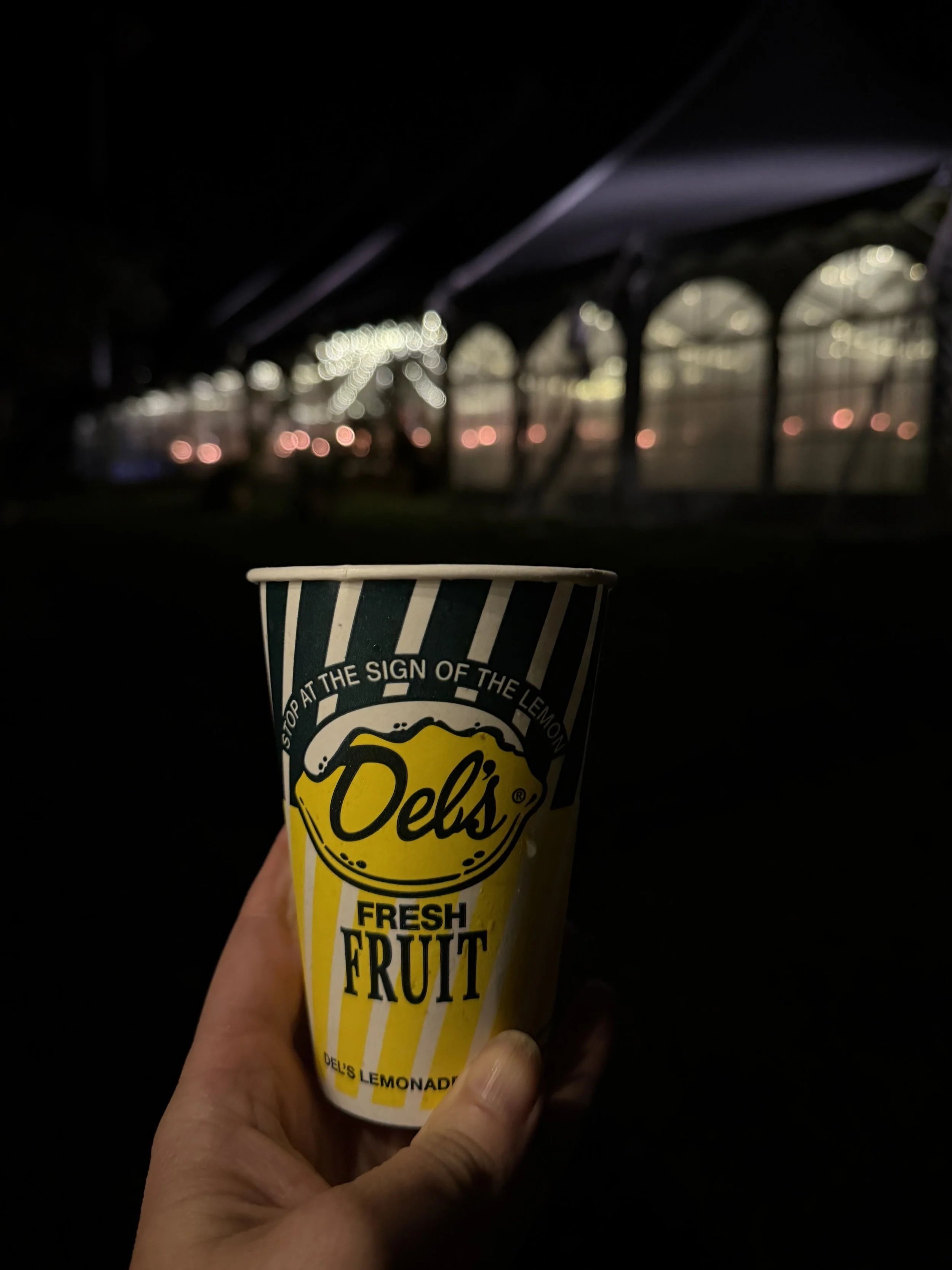 A hand holding a Del's Lemonade cup with yellow and white stripes, with a dark background showing a blurred structure with windows and lights.