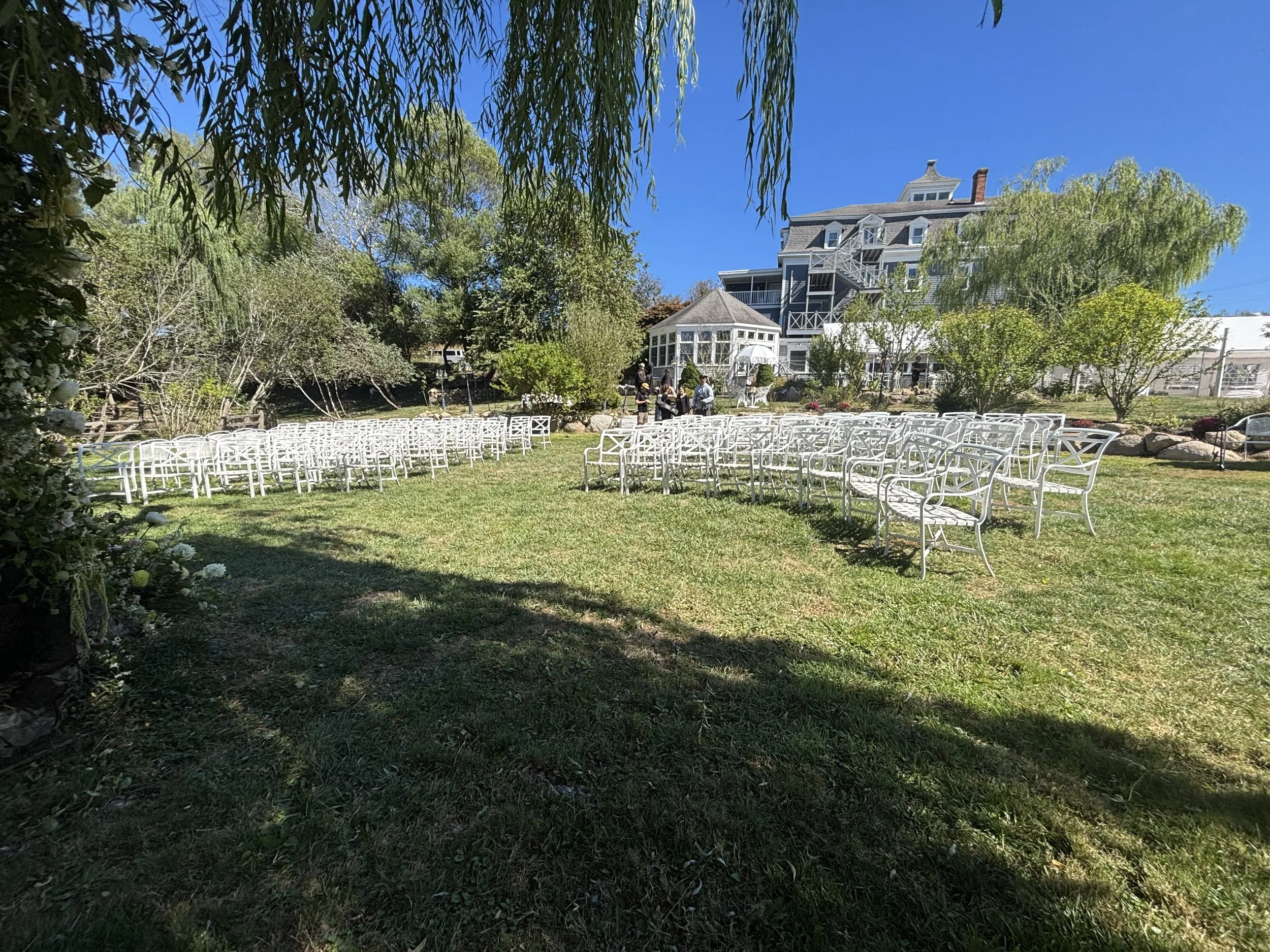 Outdoor wedding setup in a garden with rows of white chairs on the grass, a large house in the background, and several people gathered near the house under trees. The sky is clear and blue.