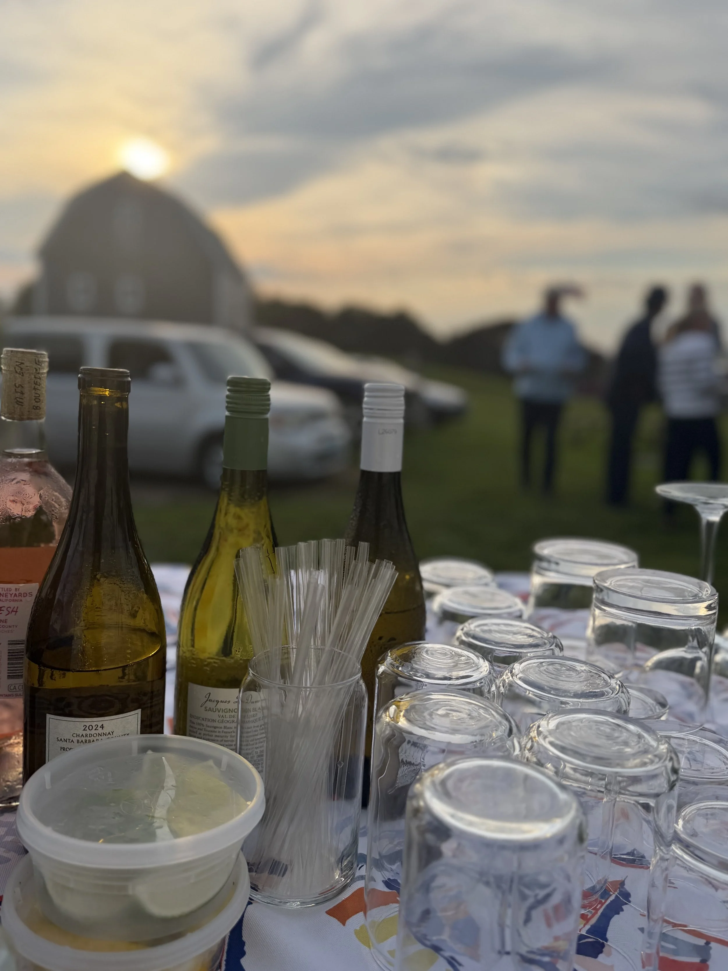 Multiple bottles and glasses on a table with people in the background outdoors during sunset.
