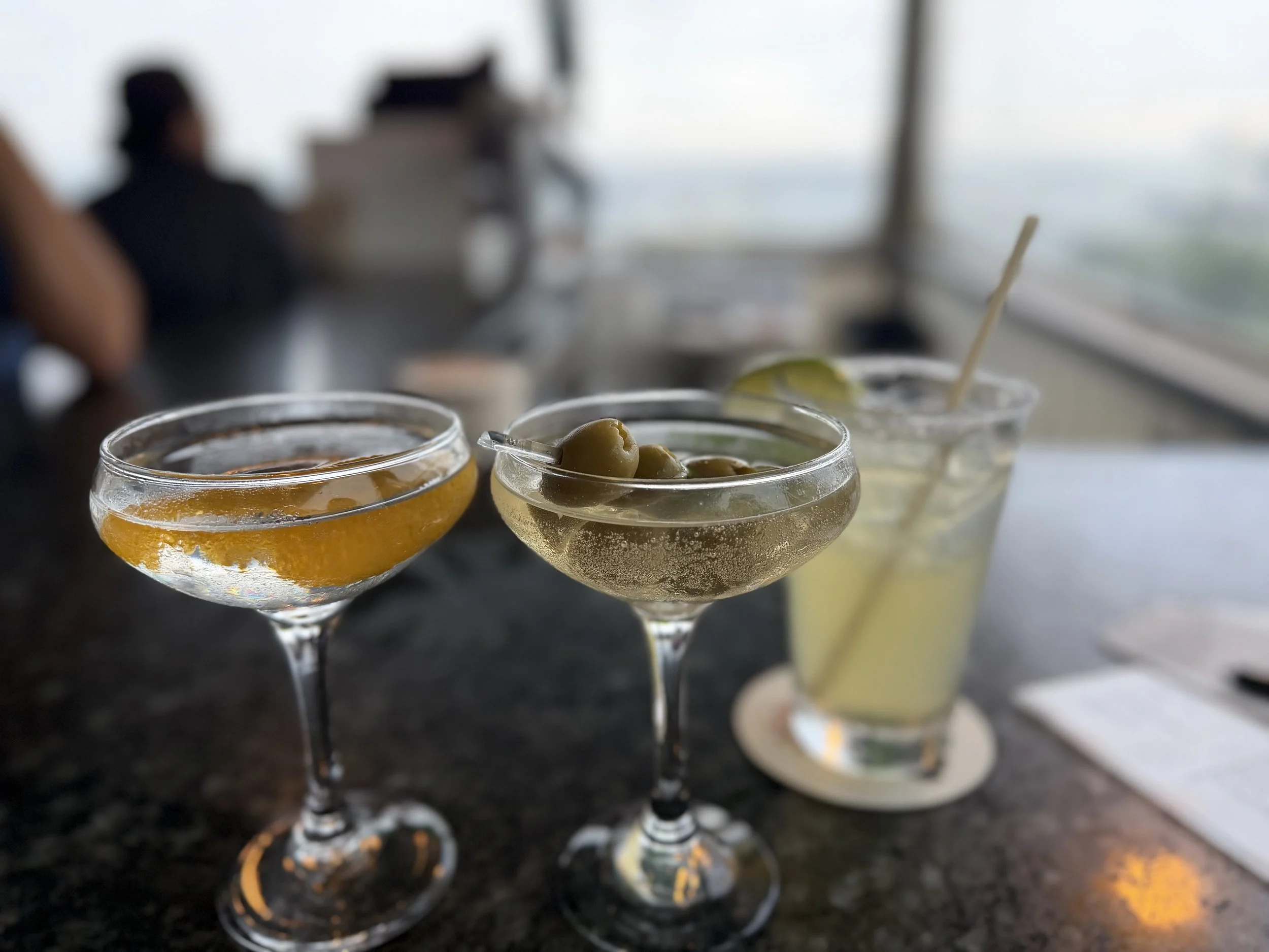 Close-up view of three cocktails in coupe glasses with olives, orange peel, and lemon slice garnish on a black countertop.