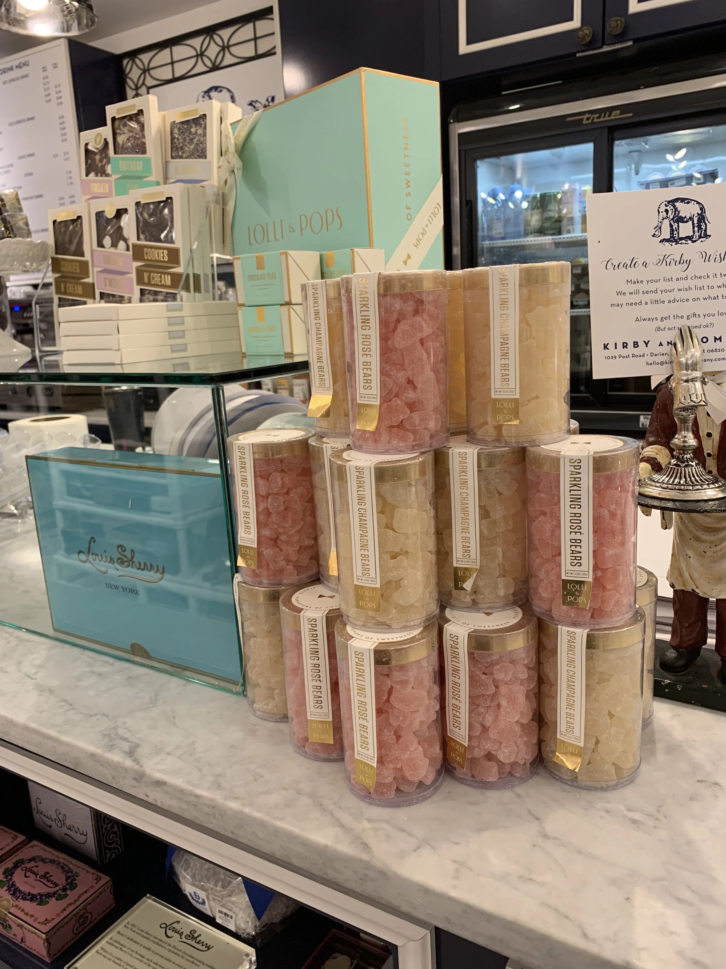 Display of pastel-colored rock candies labeled with their flavors, such as sparkling champagne, rose bears, and lemon, on a store counter with a Louis Sherry sign, in a retail environment.