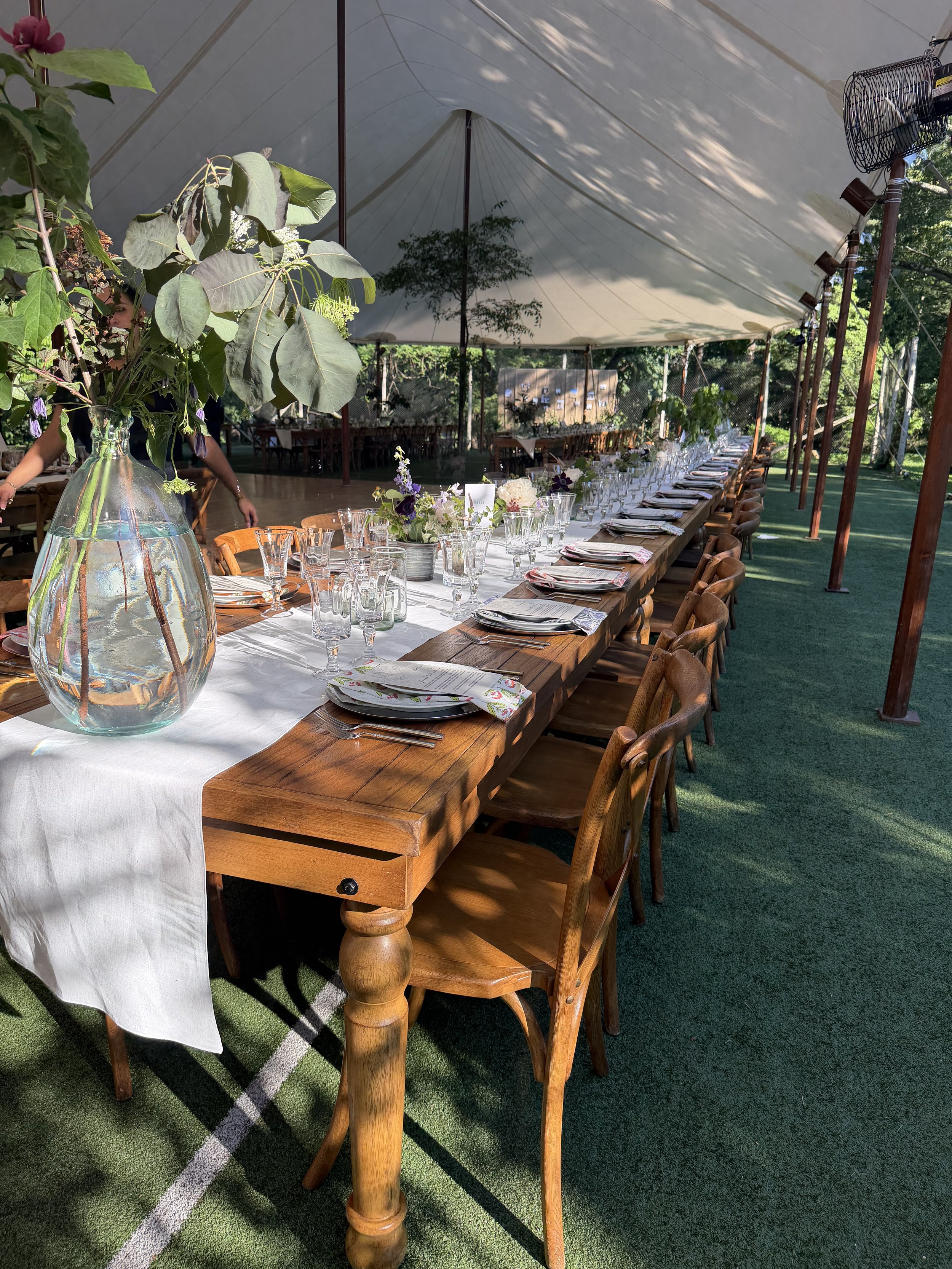 A long wooden dining table set for a formal dinner under a large white tent with greenery outside. The table is decorated with a white tablecloth, floral centerpieces, and glassware. Wooden chairs surround the table, and the area is shaded by trees.