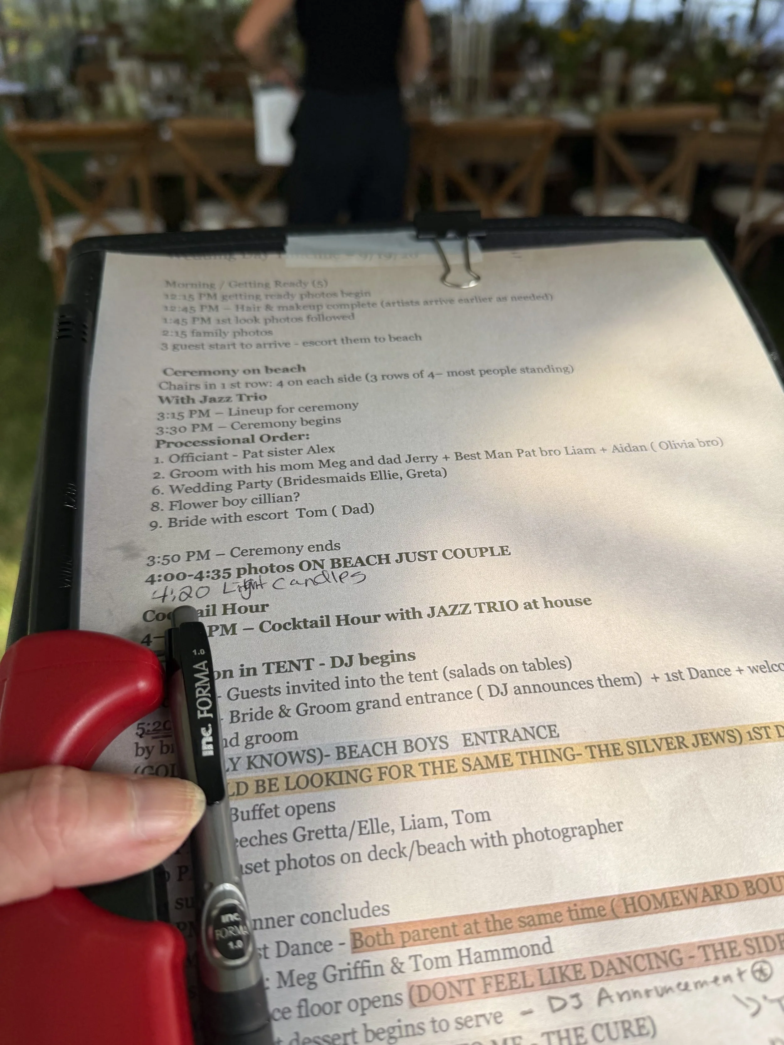 A person holding a black marker in front of a printed wedding schedule, with a blurred background of a person standing at a buffet table and chairs outdoors.