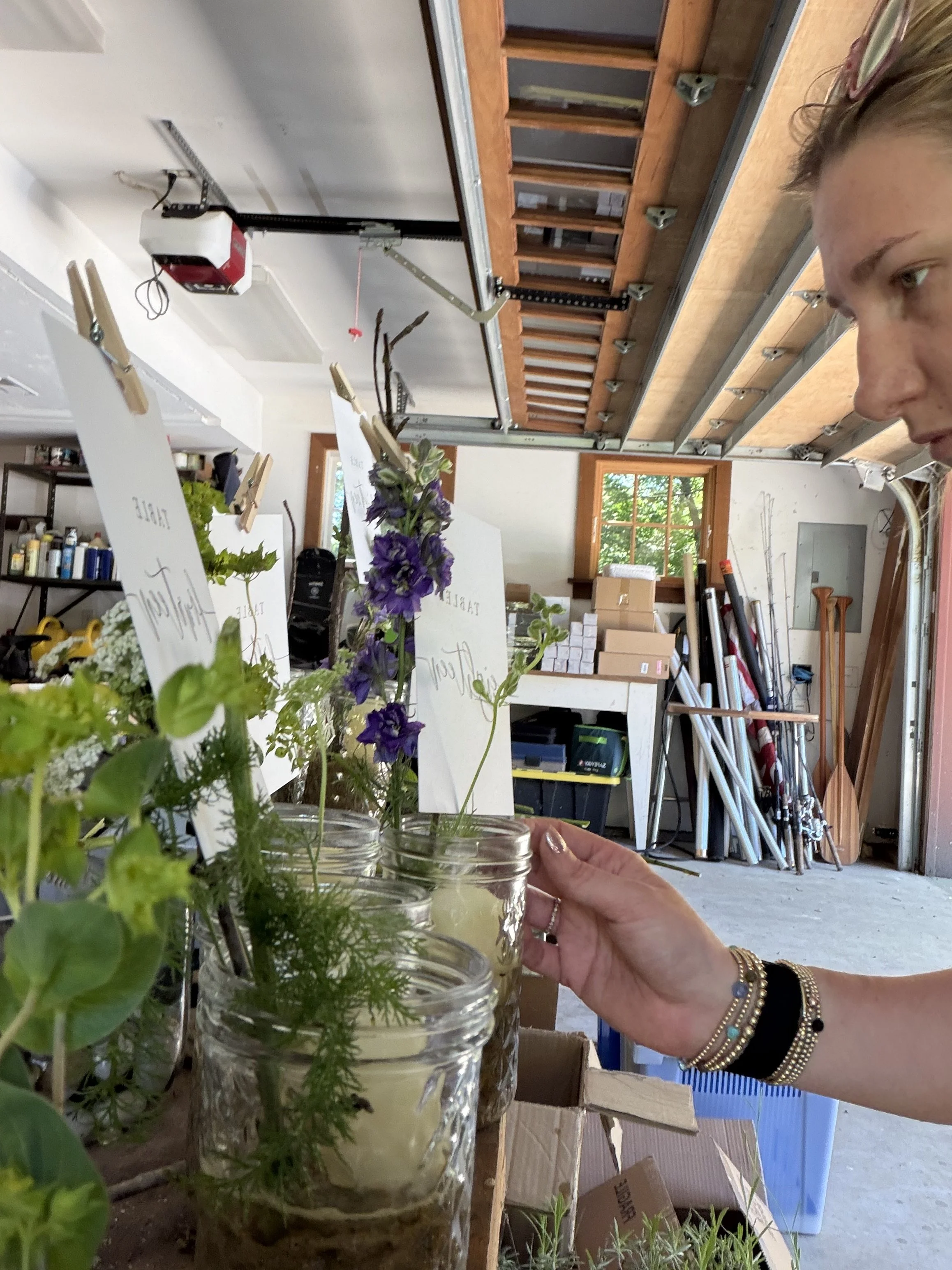 Close-up of hands arranging small potted flowers with handwritten labels in a workshop or garage, with gardening tools and supplies in the background.